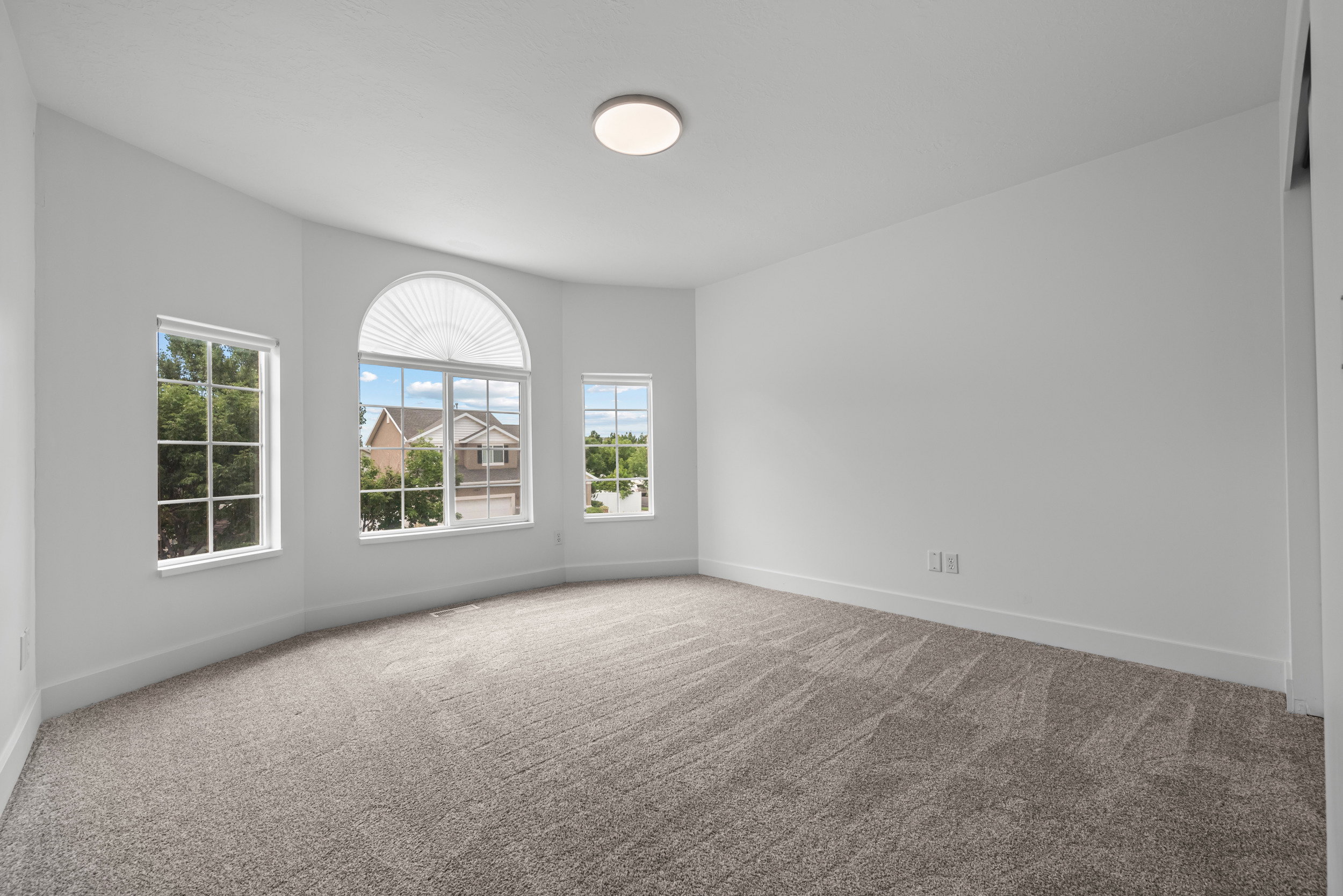 Bright room with gray carpet flooring and white walls, featuring a bay window configuration with a prominent arched window, two rectangular windows, and a flush-mount ceiling light fixture