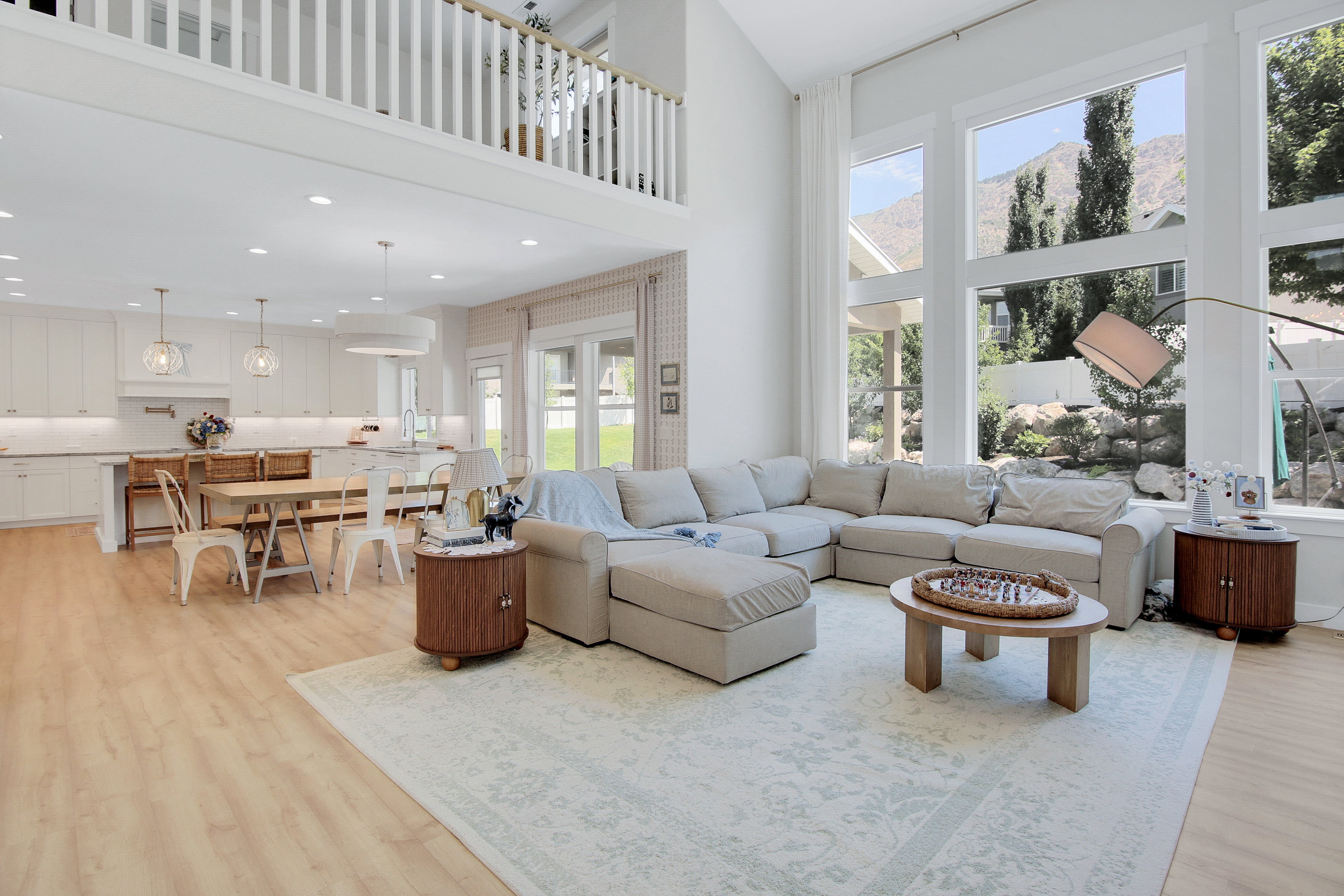 Expansive living area with high ceilings, featuring a two-story window array with mountain scenery, wood-finish flooring, and a kitchen with white cabinetry and pendant lighting