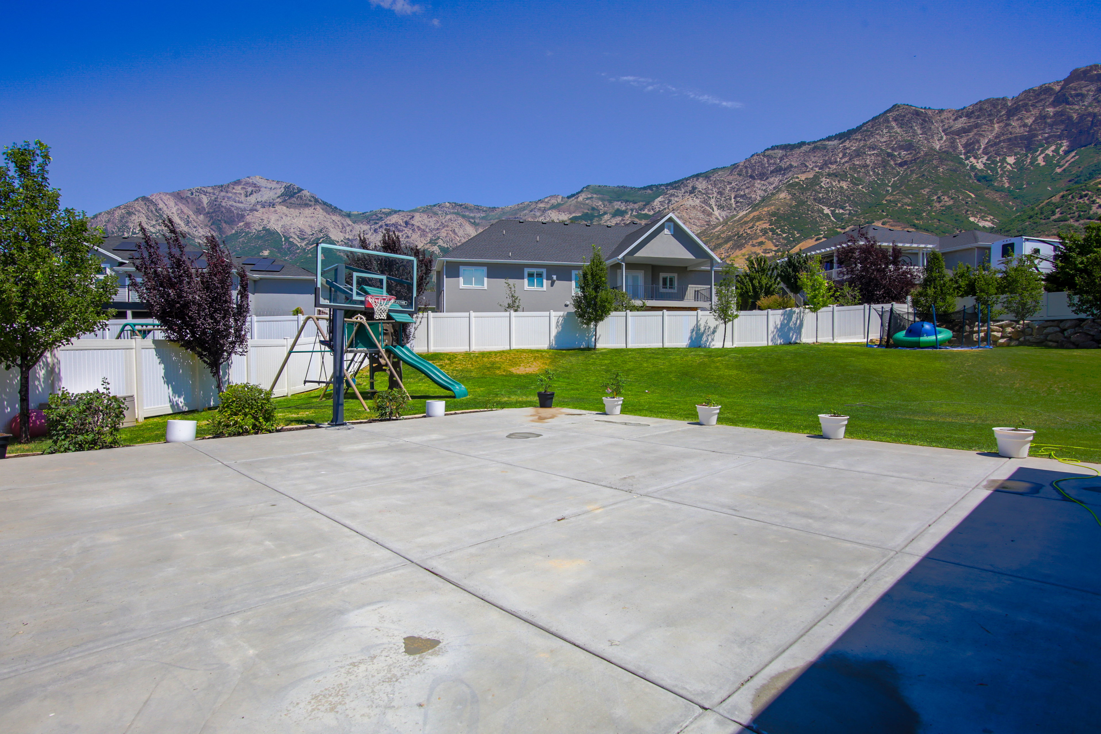 Expansive concrete patio area with a basketball hoop, bordered by a manicured lawn and white vinyl fencing