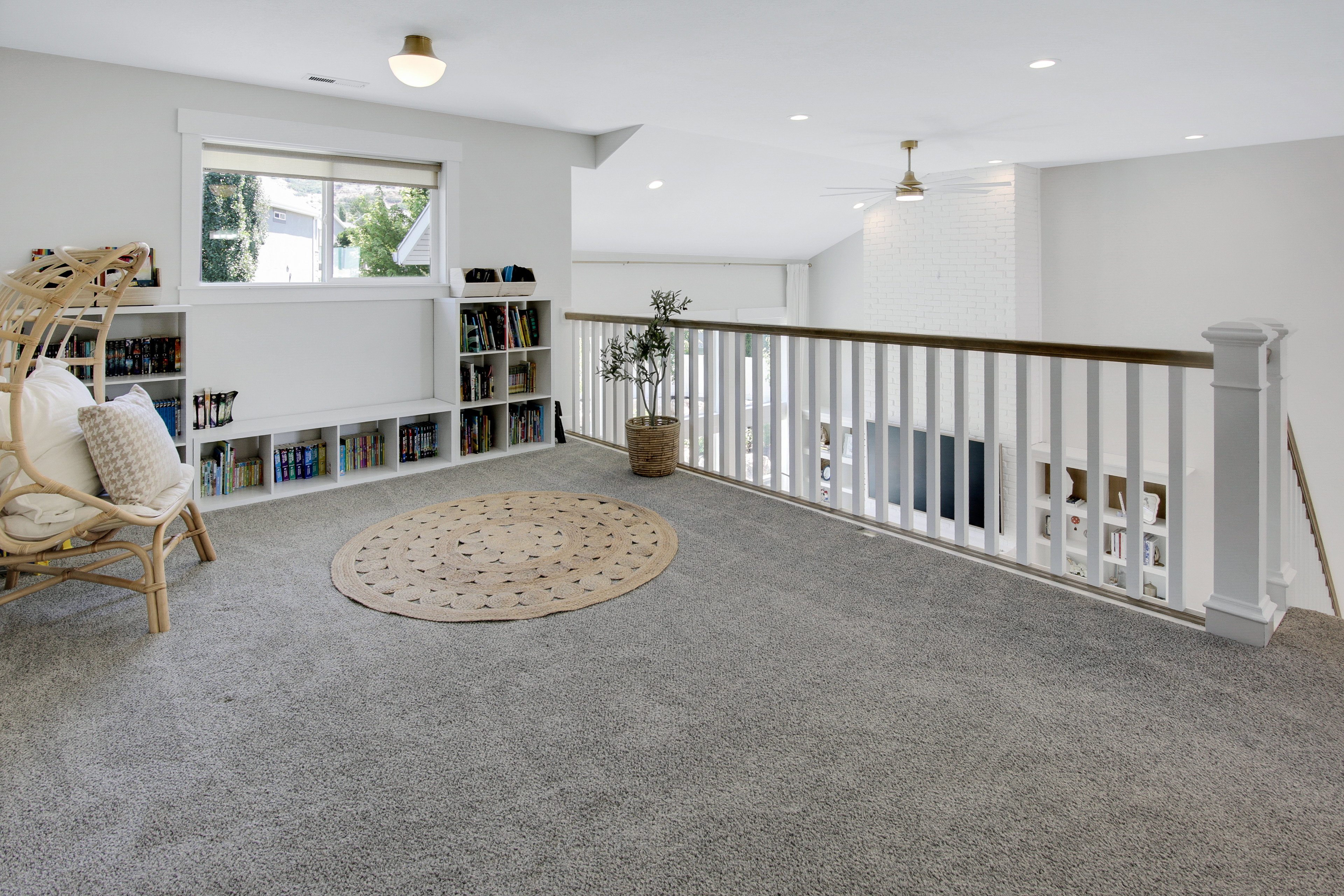 Carpeted loft area featuring a window with a white frame, built-in shelving, a white spindle railing with a wood-finish handrail, and an overhead light fixture