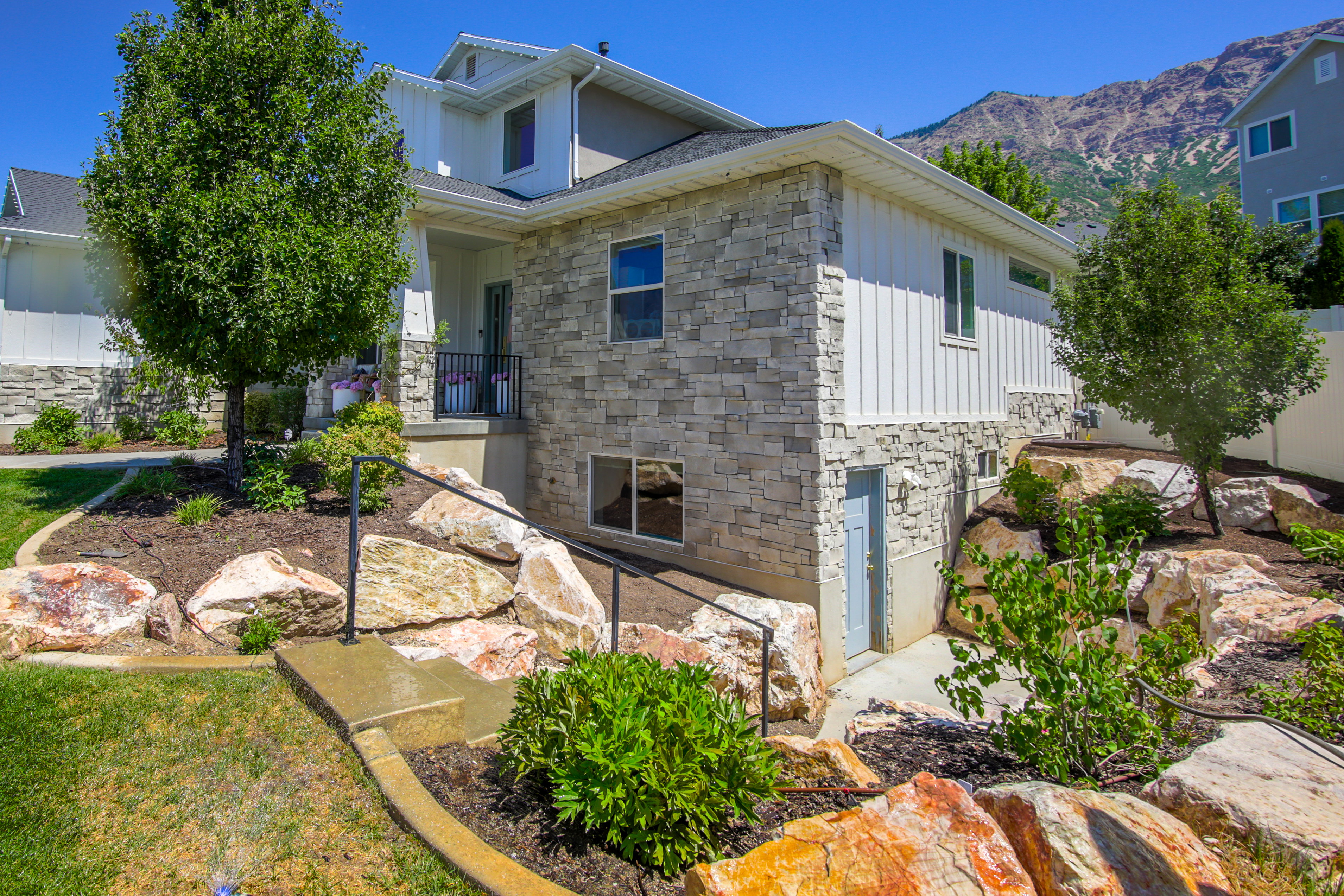 Contemporary residence featuring a stone and vertical siding exterior, a front porch with black railing, terraced landscaping with mature trees and large boulders, a concrete walkway with handrails, and a lower-level access door