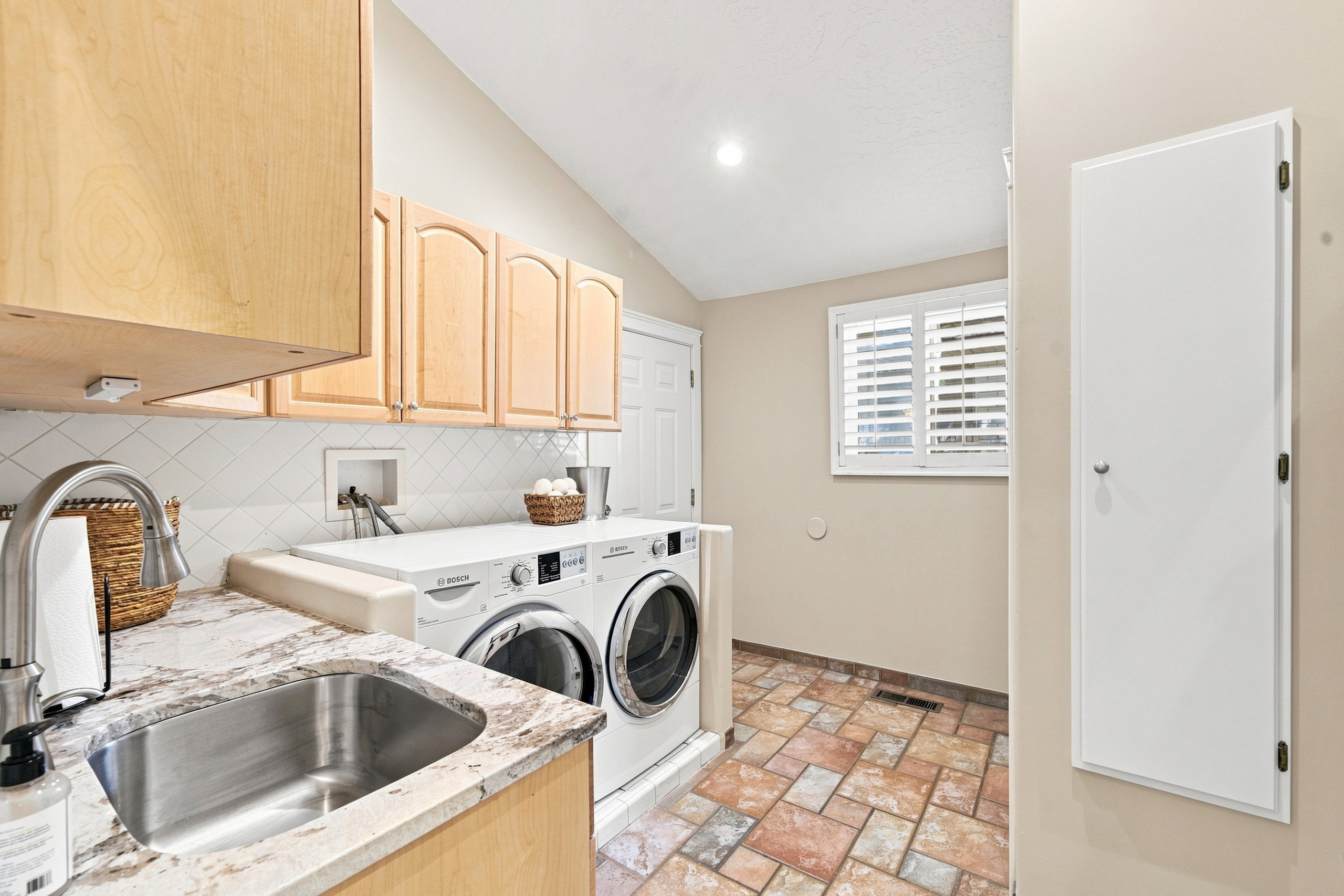 Laundry room featuring a vaulted ceiling with recessed lighting, light wood-finish cabinetry, a stainless steel sink with a high-arc faucet, a light-toned countertop, and patterned tile flooring