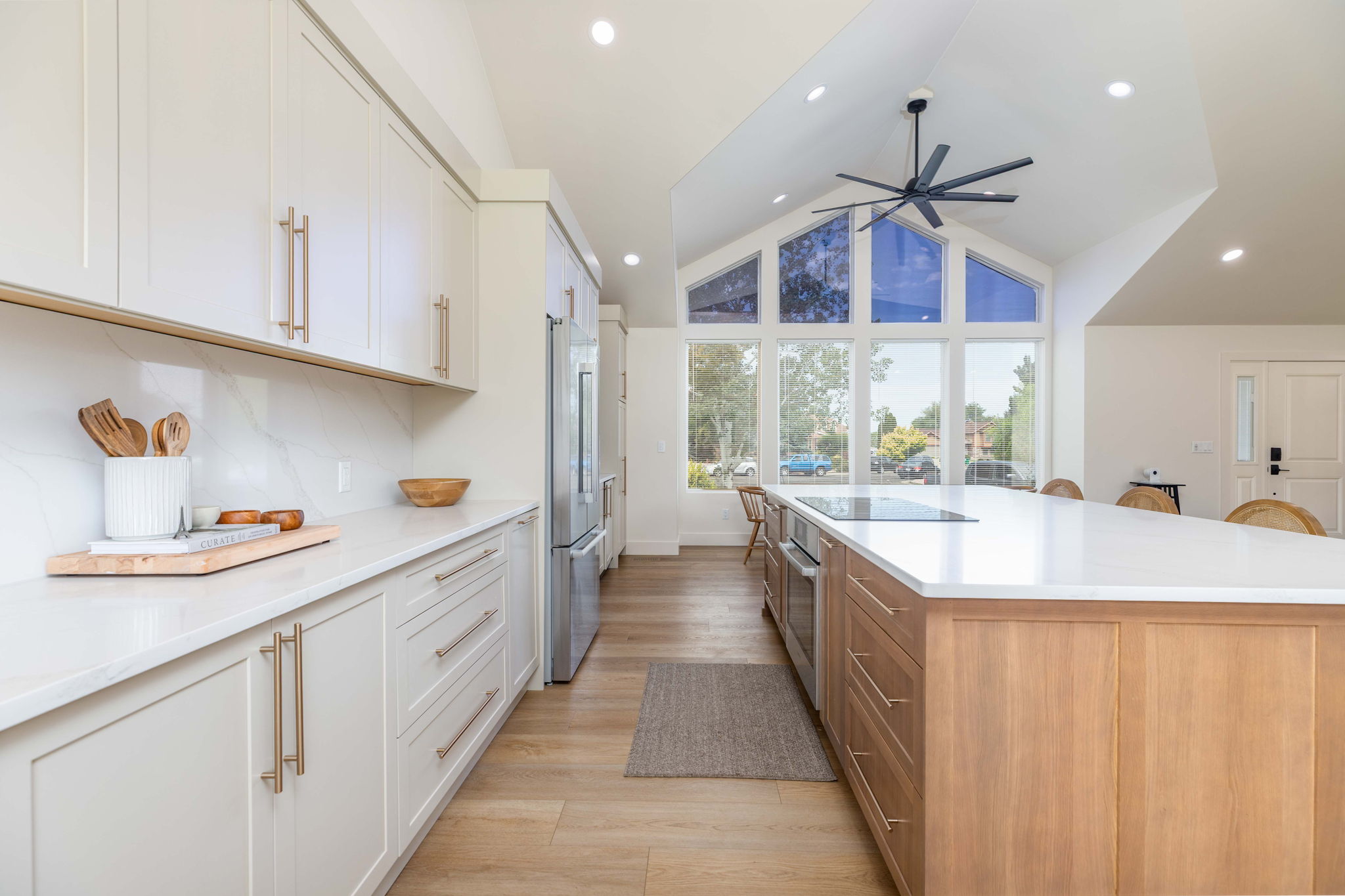 Vaulted ceiling kitchen featuring a large central island with integrated cooktop, light wood-finish flooring, full-height white cabinetry with gold-tone hardware, and a window wall with expansive exterior views