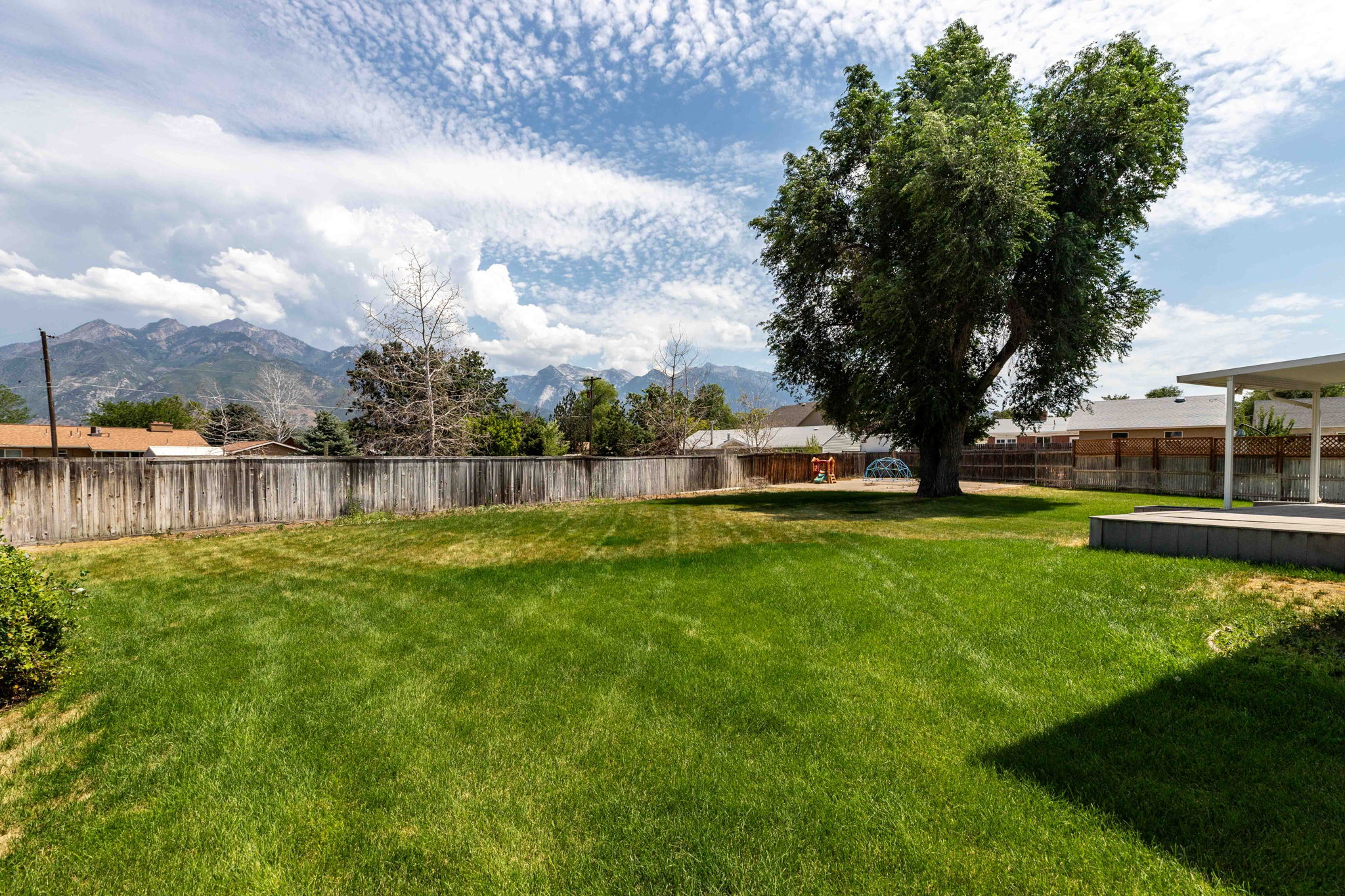 Expansive fenced backyard featuring a large mature tree, green lawn, a patio with an overhead cover, and mountain scenery