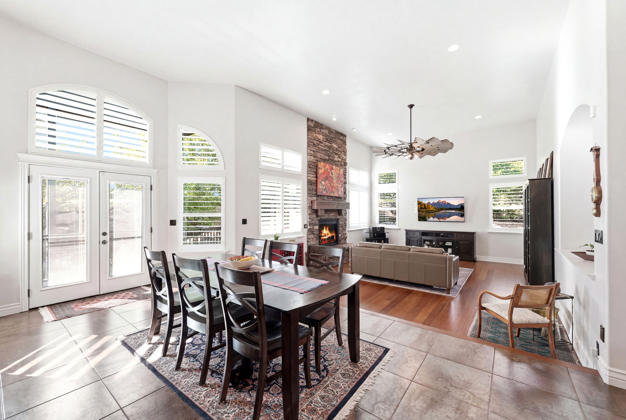 Open-concept living area with high ceilings, featuring a stone fireplace, wood-finish flooring, and multiple arched windows with plantation shutters