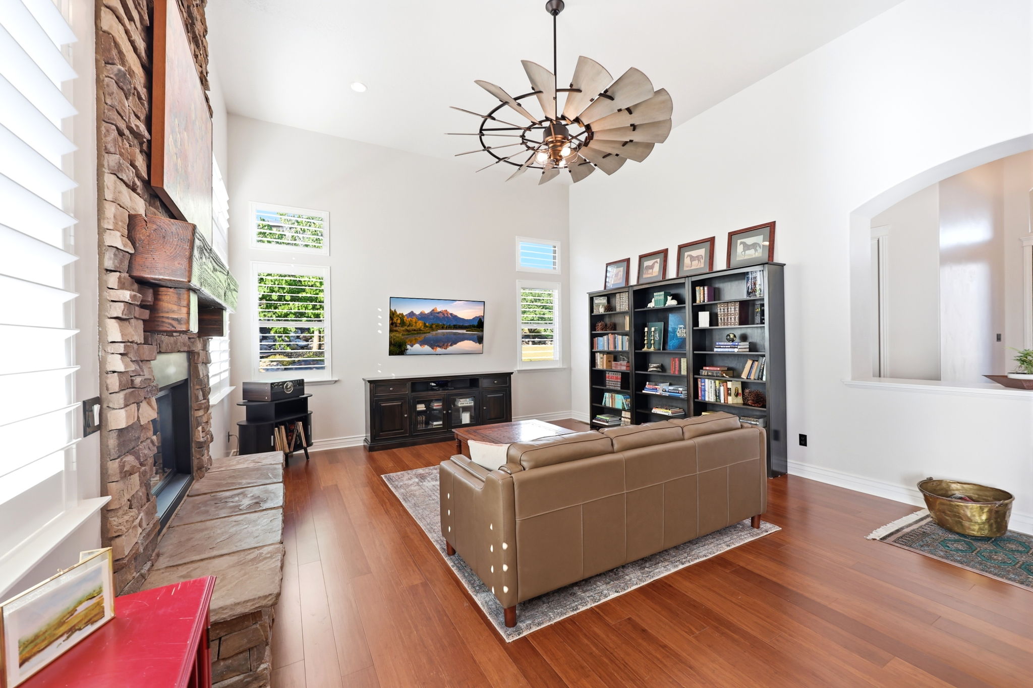 Spacious living area featuring high vaulted ceilings, wood-finish flooring, a stone-clad fireplace with a stone hearth, multiple windows with plantation shutters, and a unique ceiling fan fixture