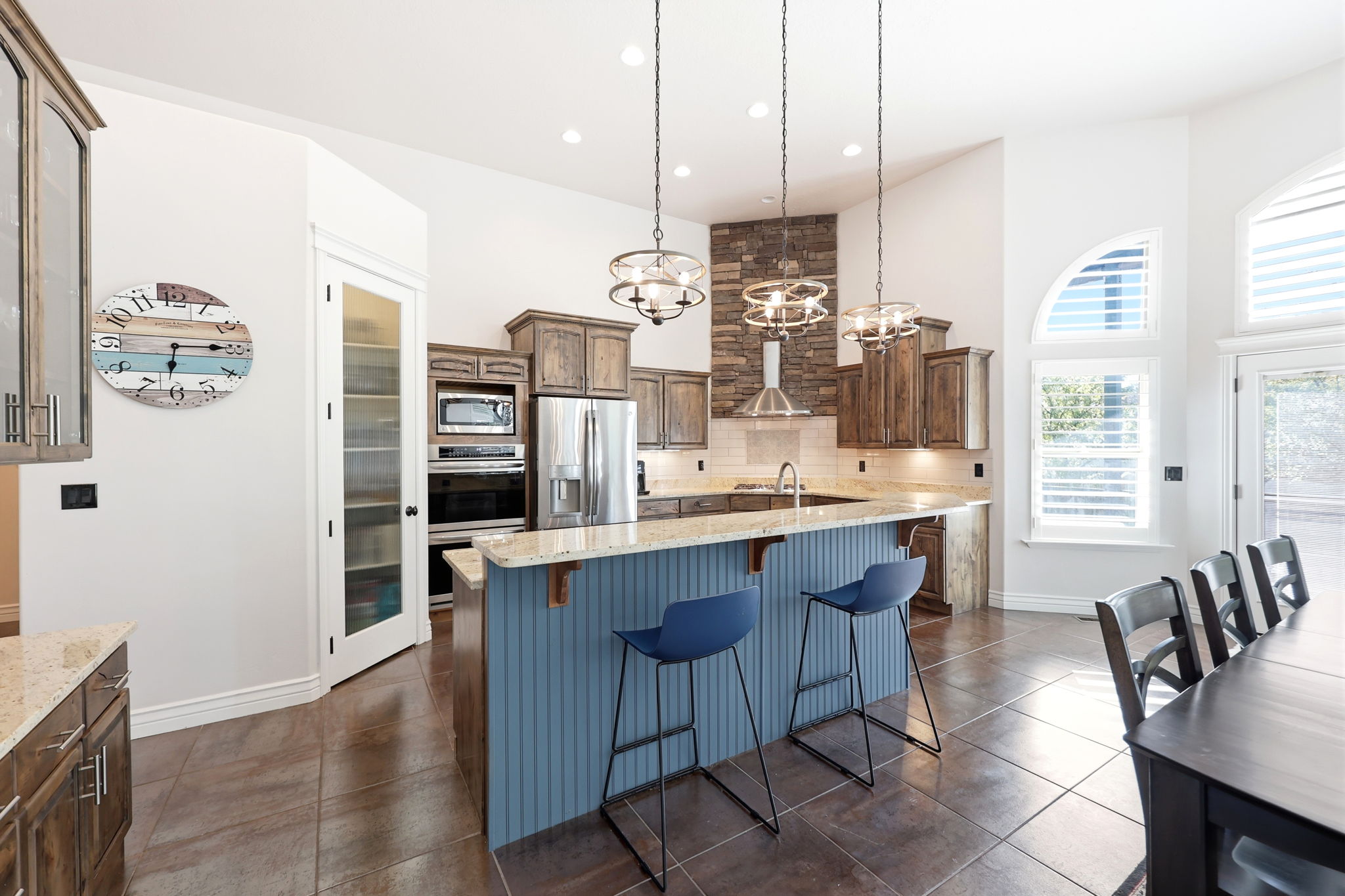 Spacious kitchen featuring a large central island with beadboard paneling, integrated stainless steel appliances, stacked stone range hood surround, wood cabinetry, and light-toned countertops