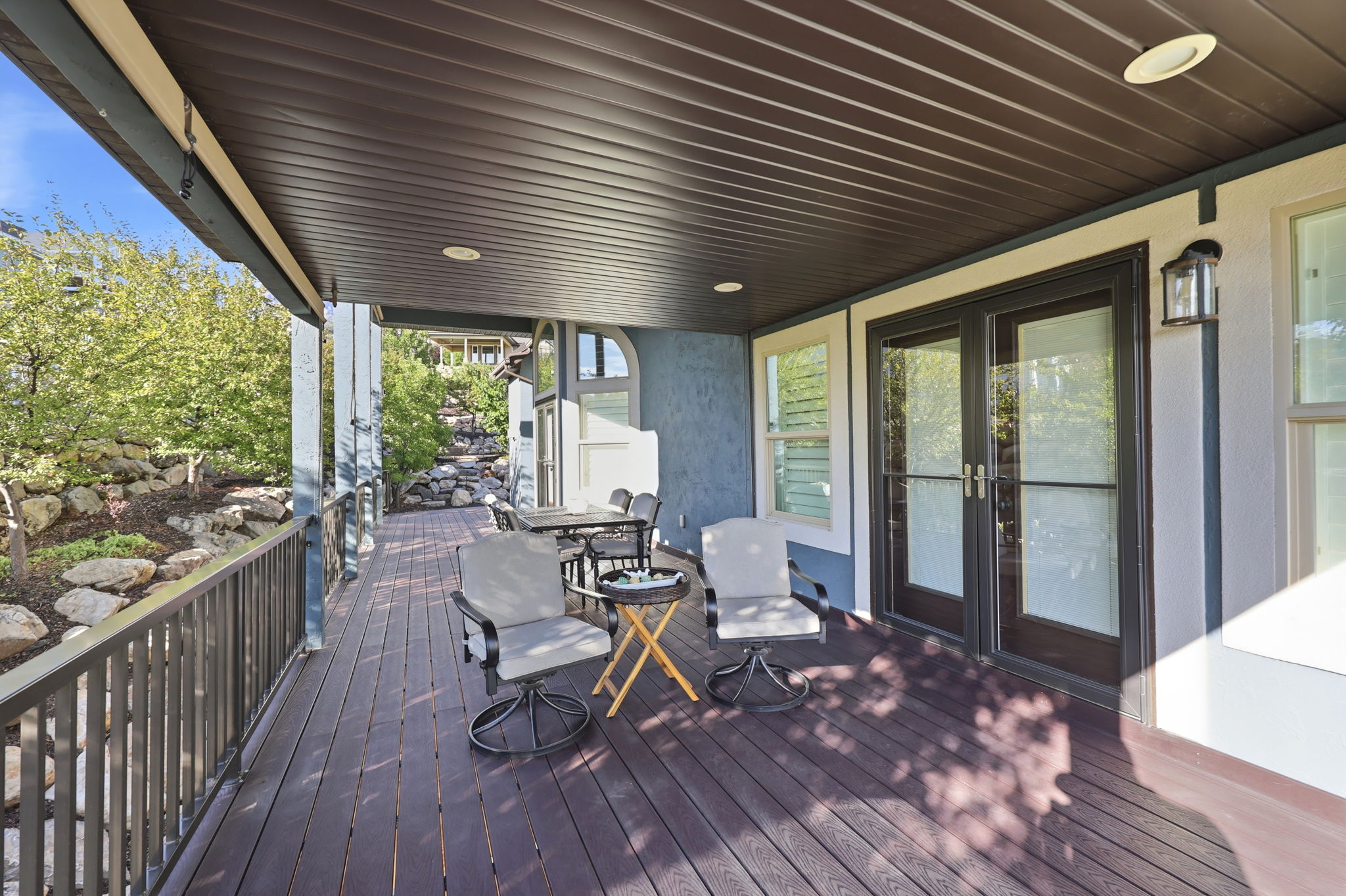 Covered outdoor deck with dark wood-finish flooring, featuring a dark metal railing, recessed lighting, and direct access through double glass doors