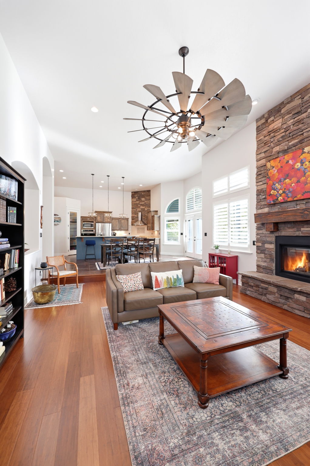 Spacious living area featuring high ceilings, wood-finish flooring, a stacked stone fireplace with a wood mantel, and multiple arched window openings with shutters