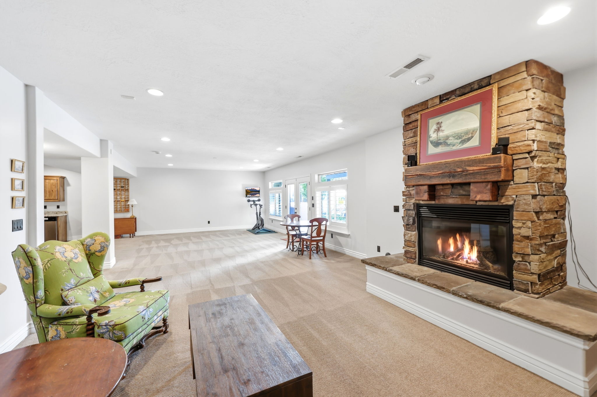 Spacious room featuring a stone fireplace with a substantial timber mantel, recessed lighting, expansive carpeted flooring, and a series of windows providing natural illumination