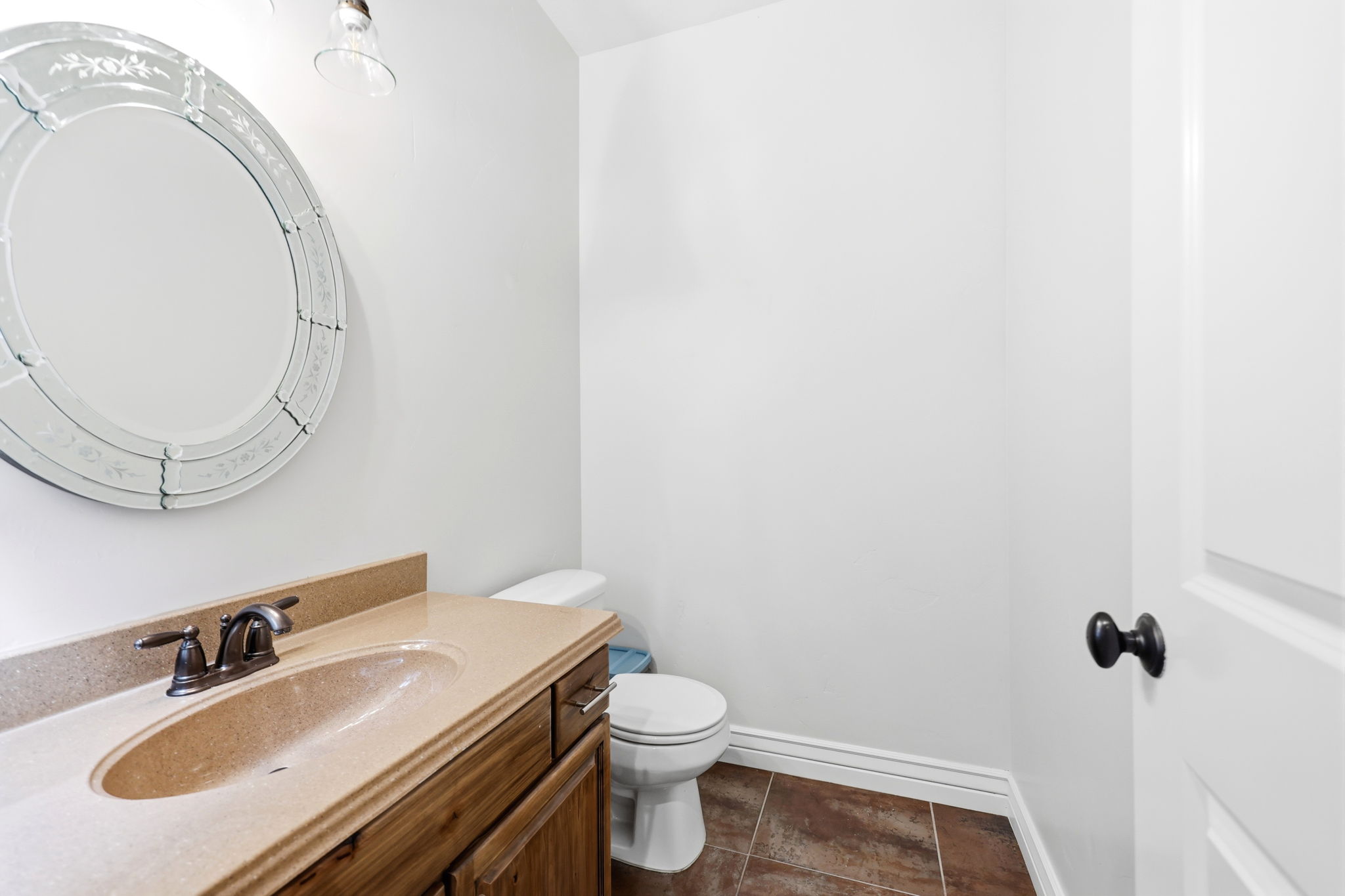 Powder room featuring a wood-finish vanity with an integrated sink and stone-look countertop, an ornate round mirror, a toilet, and tile flooring