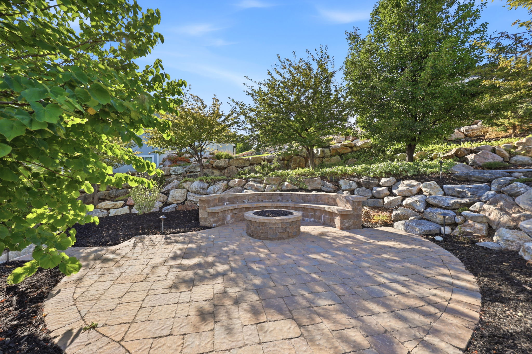 Paver patio featuring a built-in fire pit, stone seating, and extensive rock landscaping with mature trees