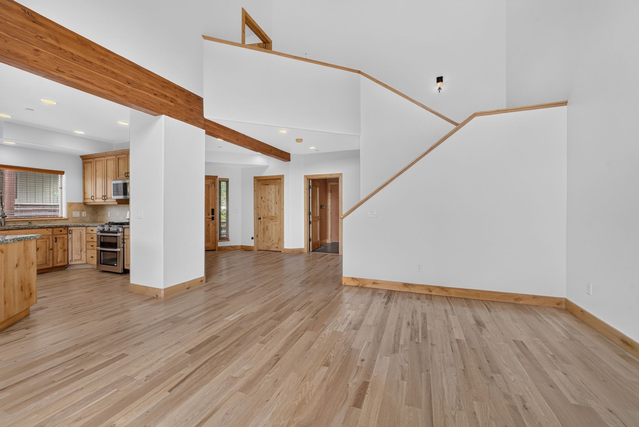 Open-concept interior featuring light wood-finish flooring, exposed wood ceiling beams, a kitchen with wood cabinetry and a stainless steel oven, and a partial staircase with wood trim