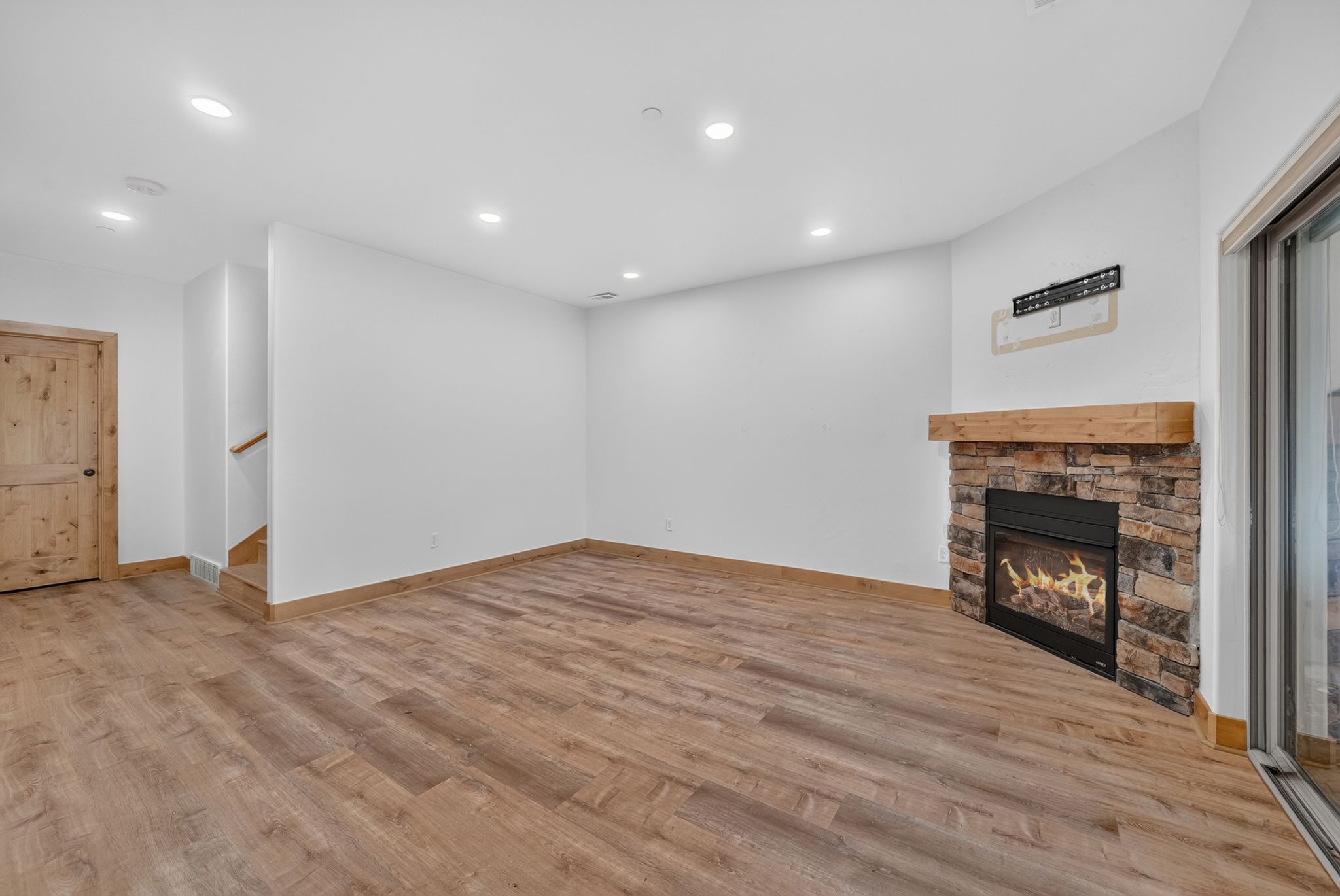 Spacious living area featuring wood-finish flooring, a stone-clad fireplace with a wood mantel, recessed lighting, and a sliding glass door