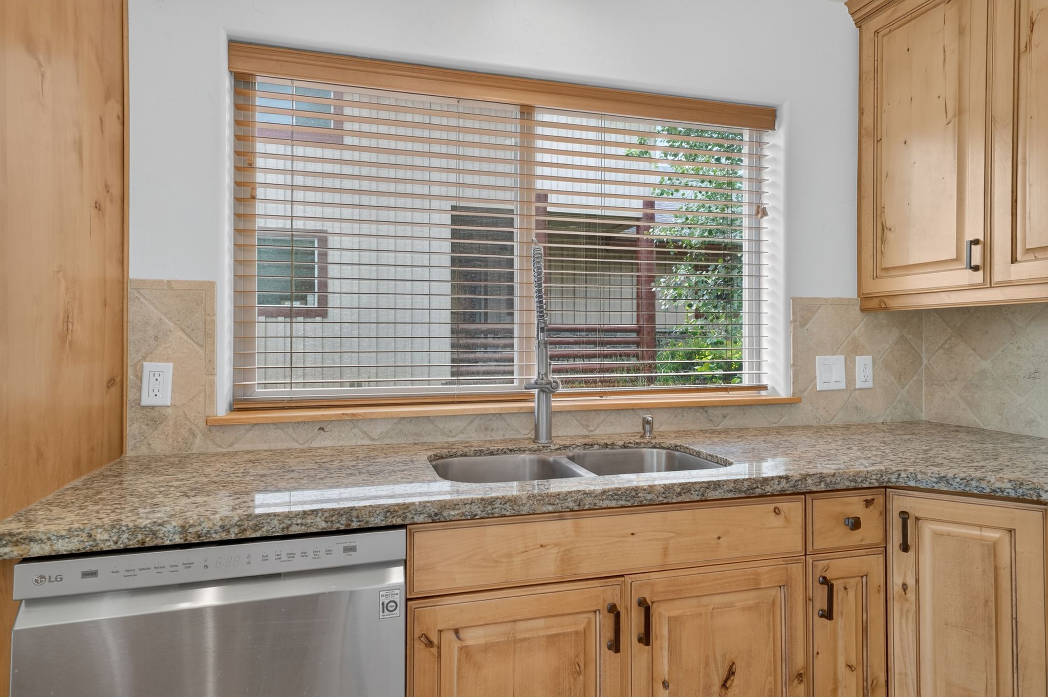 Kitchen featuring light wood cabinetry, granite-finish countertops, stainless steel dual-basin sink with a gooseneck faucet, and a tiled backsplash