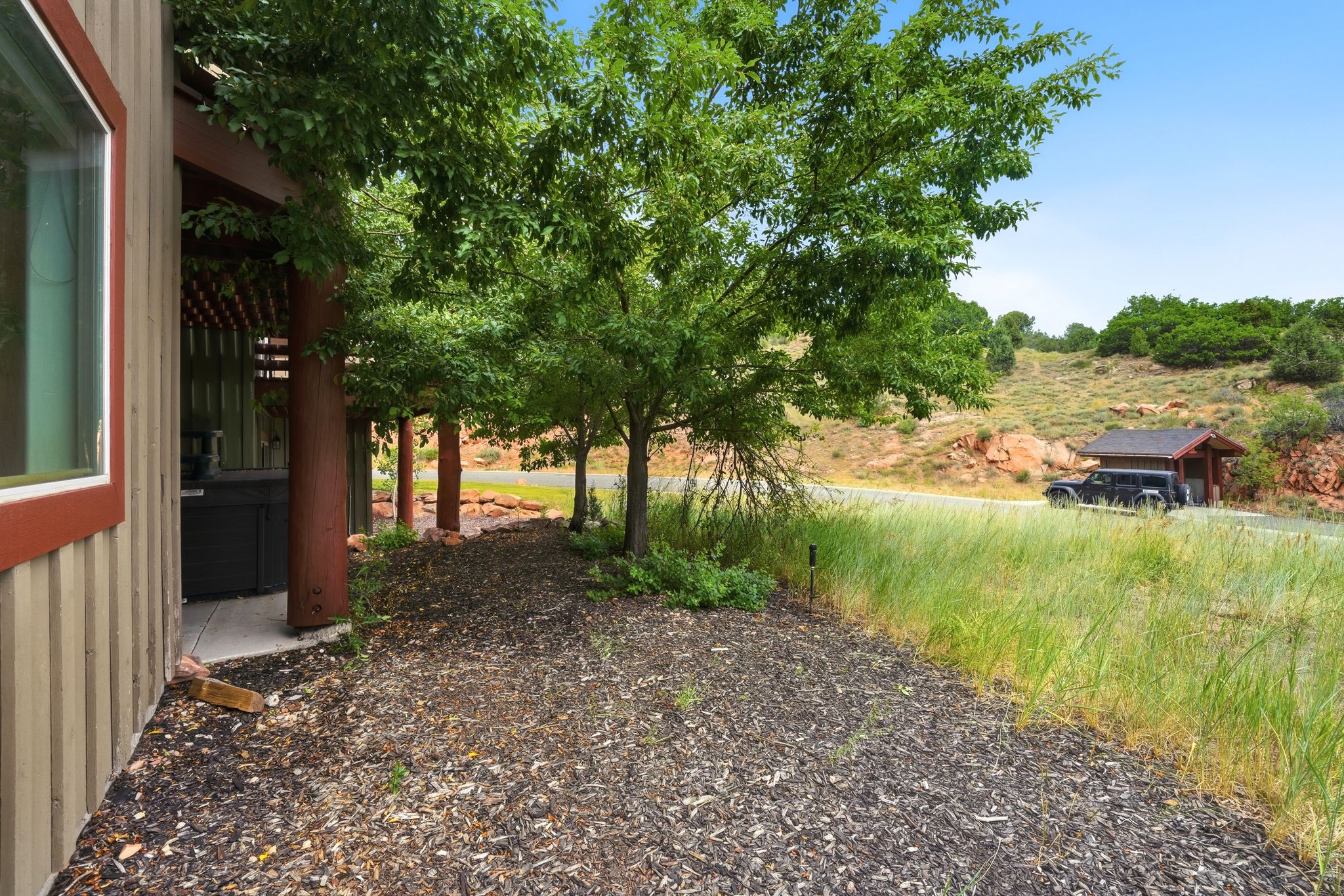 Side patio featuring a built-in hot tub under a wood pergola, wood-sided exterior, mature trees, and a gravel-mulch ground covering