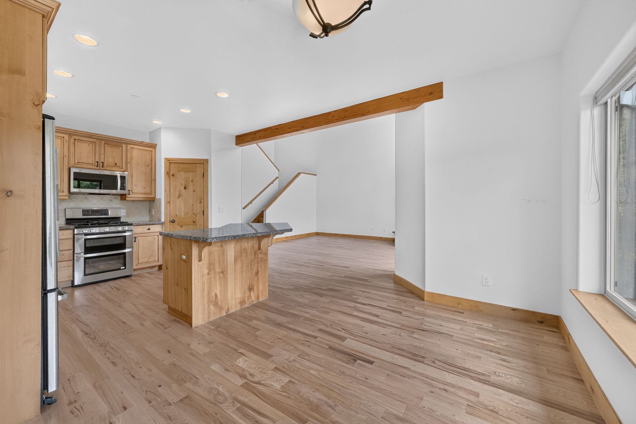 Open-concept living space featuring wood-finish flooring, a kitchen island with stone countertop, stainless steel appliances, recessed lighting, and an exposed wooden beam
