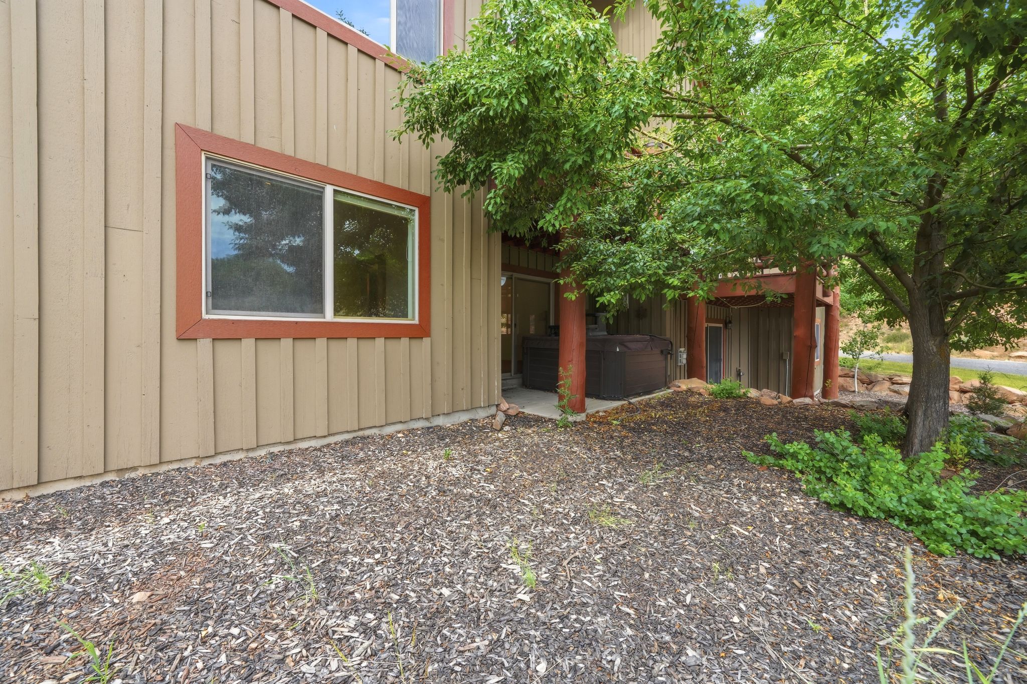 Exterior wall with tan vertical siding, a rectangular window with a red-brown frame, and an uncovered patio area with a dark spa