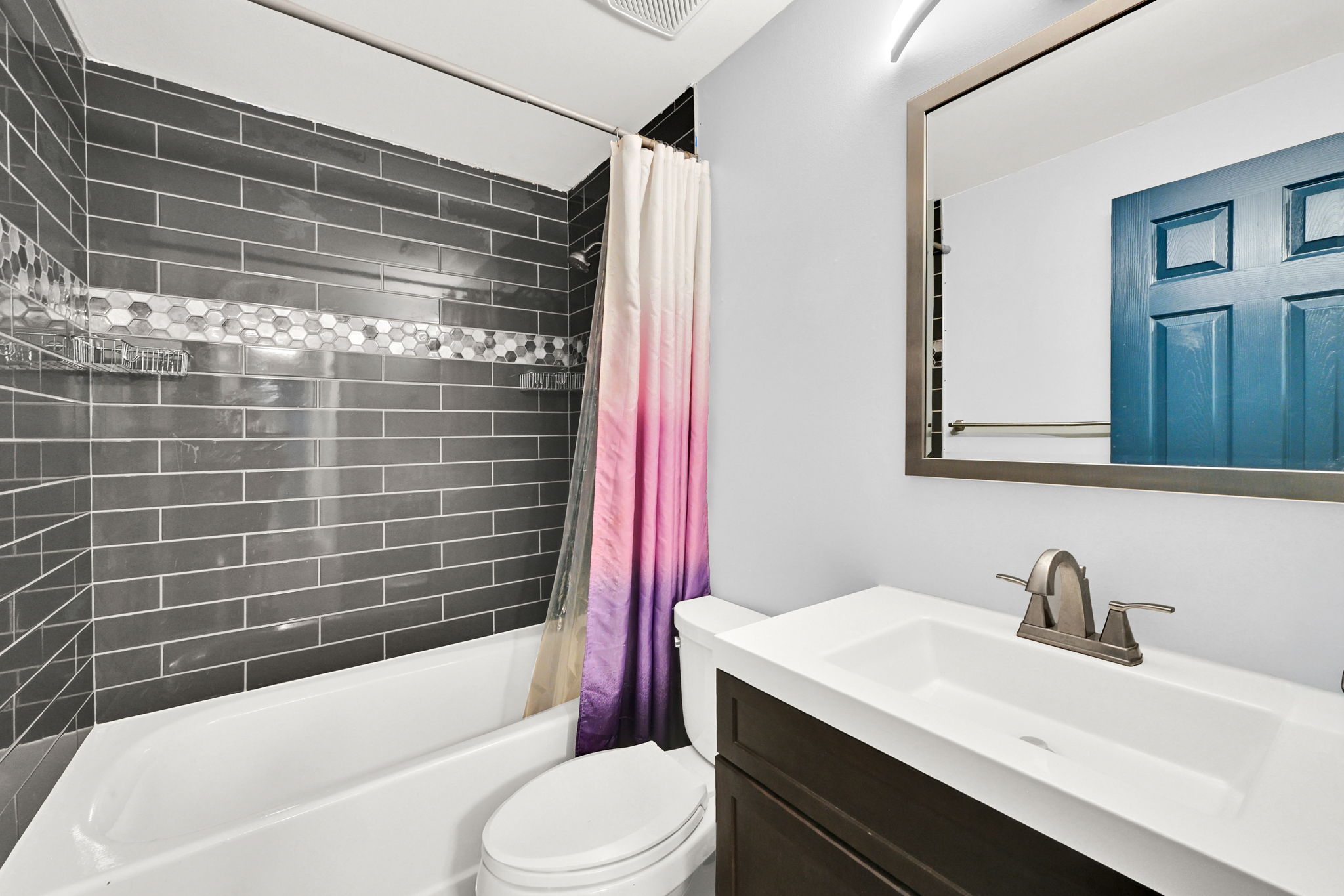 Contemporary bathroom featuring a white tub, dark grey subway tile surround with a hexagonal mosaic accent, a dark wood-finish vanity with an integrated sink, and a framed wall mirror