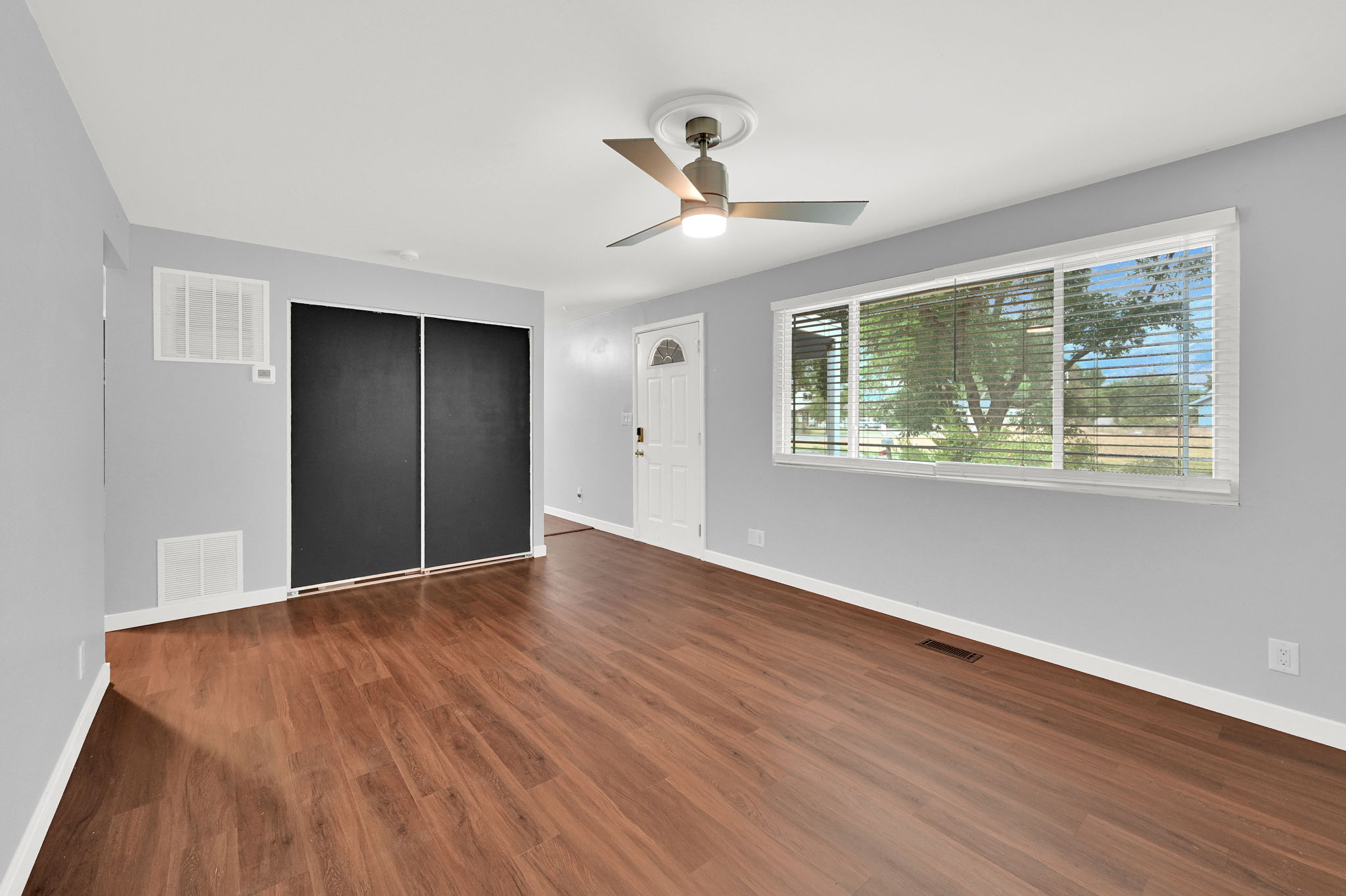 Spacious room featuring wood-finish flooring, light grey walls, and a large window with white blinds