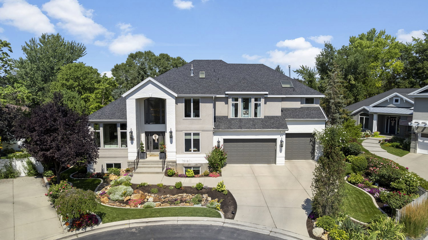 Contemporary residence featuring a prominent gabled entry with an arched window, a dark shingled roof, a paved driveway leading to a three-car garage, and extensive landscaping with rock features and mature shrubbery