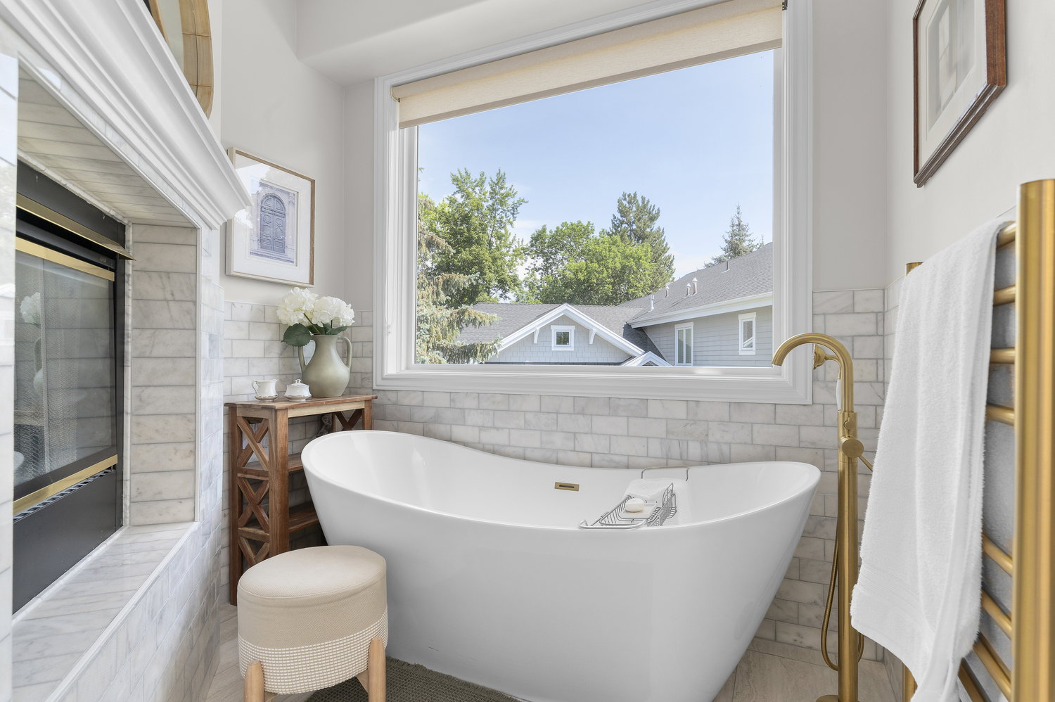 Freestanding soaking tub with a floor-mounted gold-finish faucet, subway tile surround, built-in fireplace, and large window