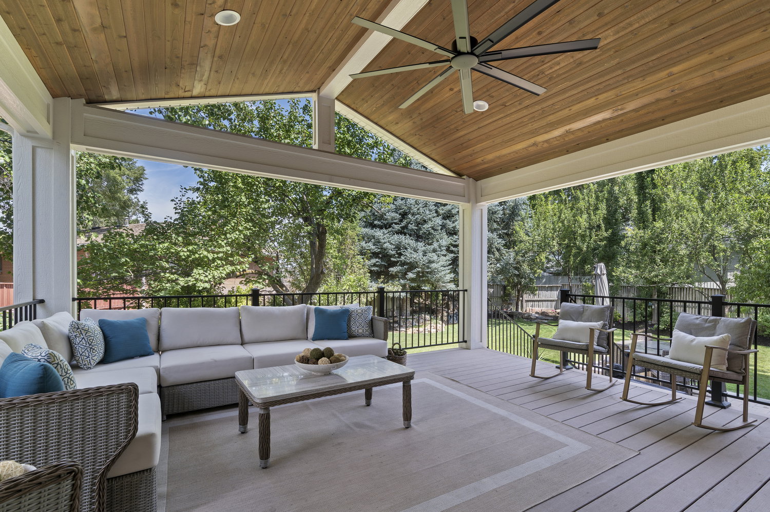 Spacious covered outdoor living area featuring a vaulted wood-finish ceiling with recessed lighting and a ceiling fan, composite decking, and black metal railings overlooking a landscaped yard