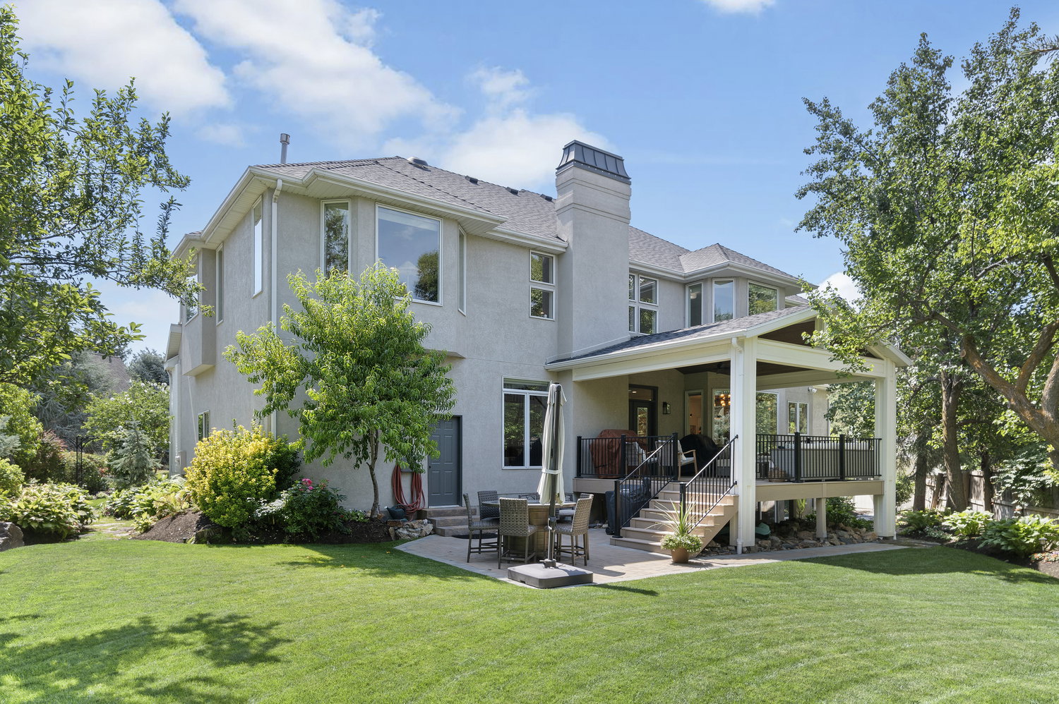 Expansive rear exterior featuring a covered elevated patio with dark railings, a lower-level concrete patio, a stucco exterior, and a spacious lawn