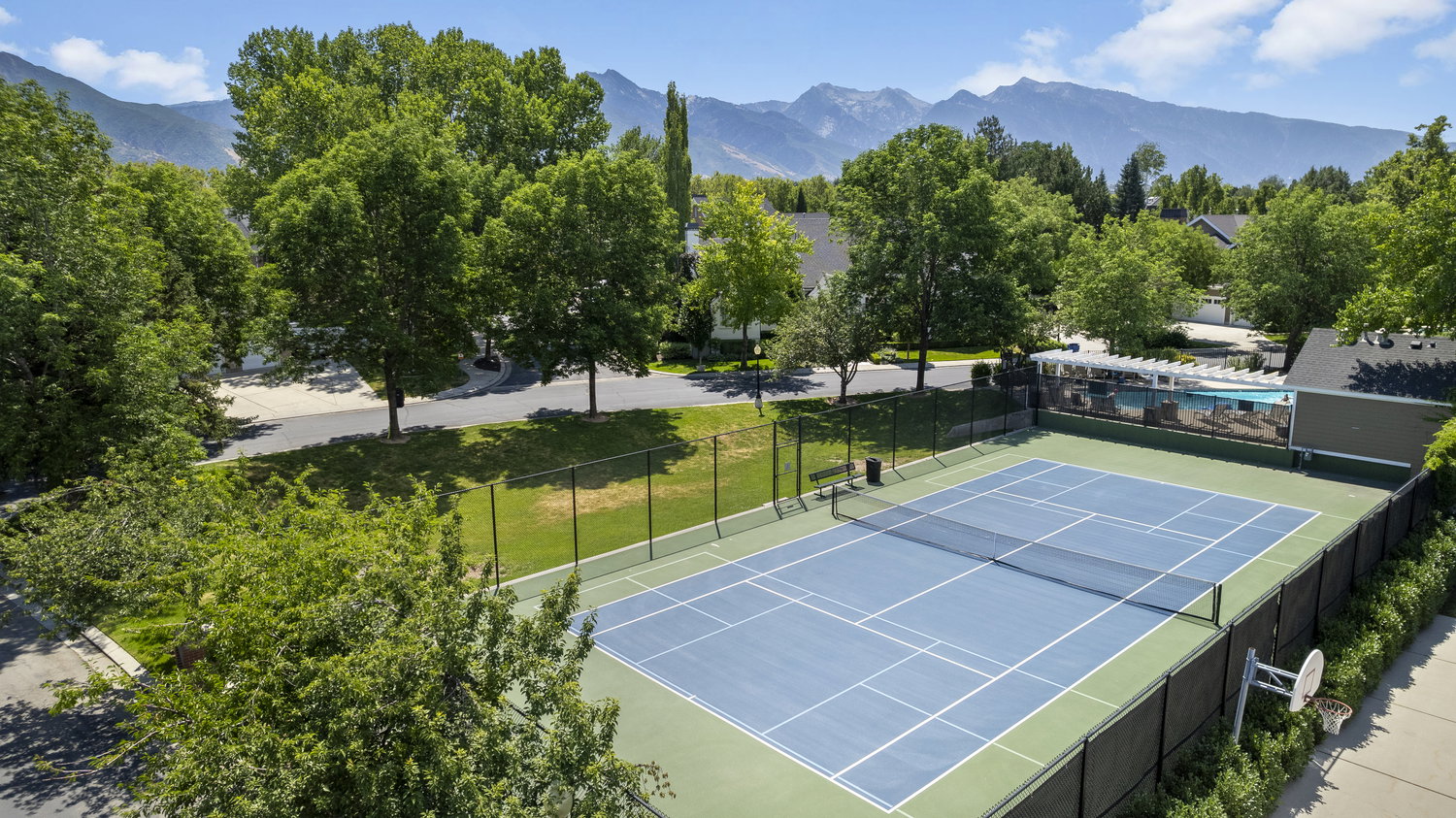 Full-size tennis court featuring a blue playing surface and green surrounds, enclosed by a black chain-link fence