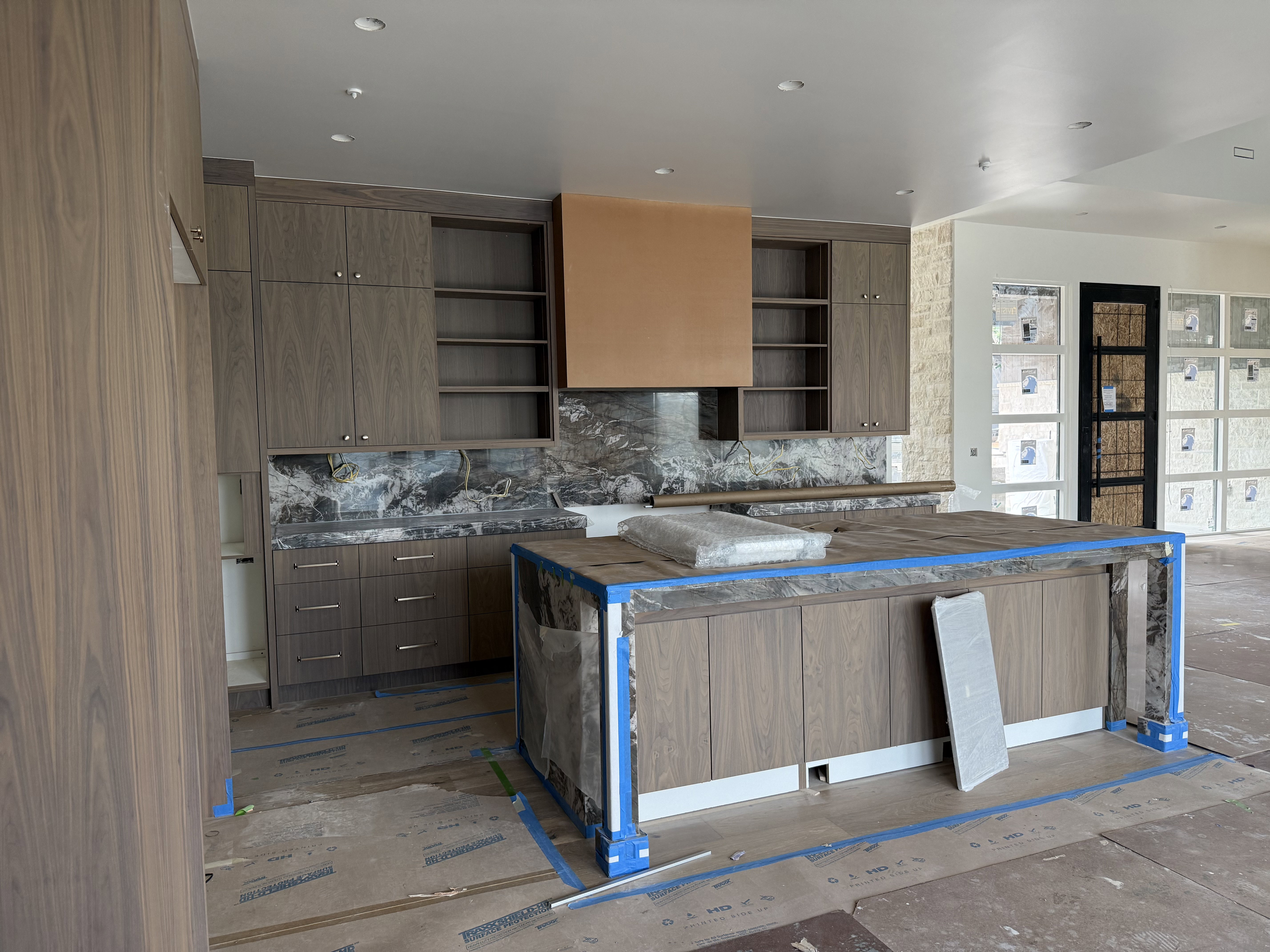 Contemporary kitchen featuring wood-finish cabinetry, a large center island with stone countertop, integrated appliances, a decorative range hood, and a stone slab backsplash