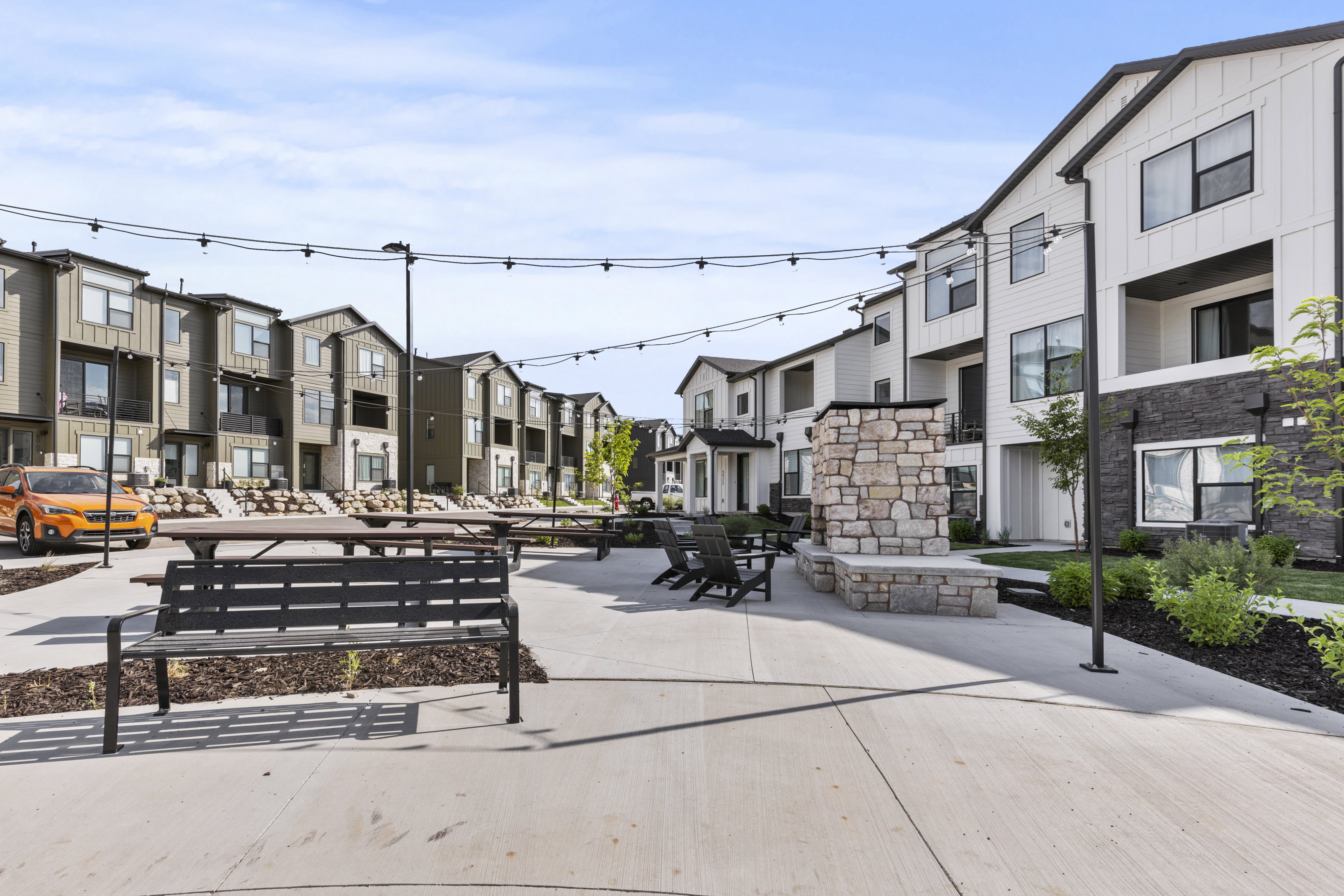 Community courtyard featuring concrete paving, a stone-clad outdoor fireplace, string lighting, and ample seating areas with picnic tables and benches