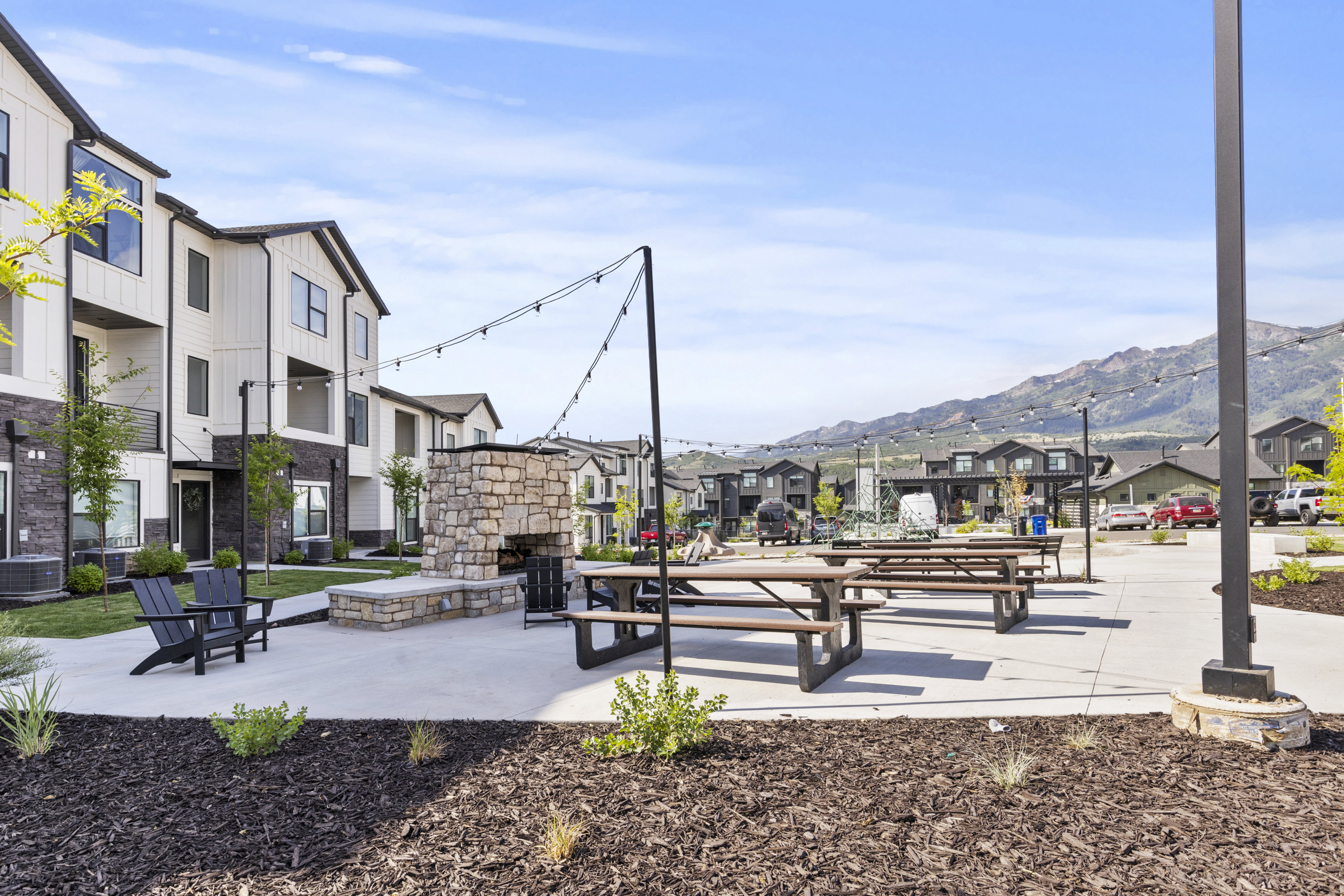 Outdoor common area featuring a stone fireplace, multiple picnic tables, strung overhead lighting, and concrete paving, surrounded by white and black multi-story buildings