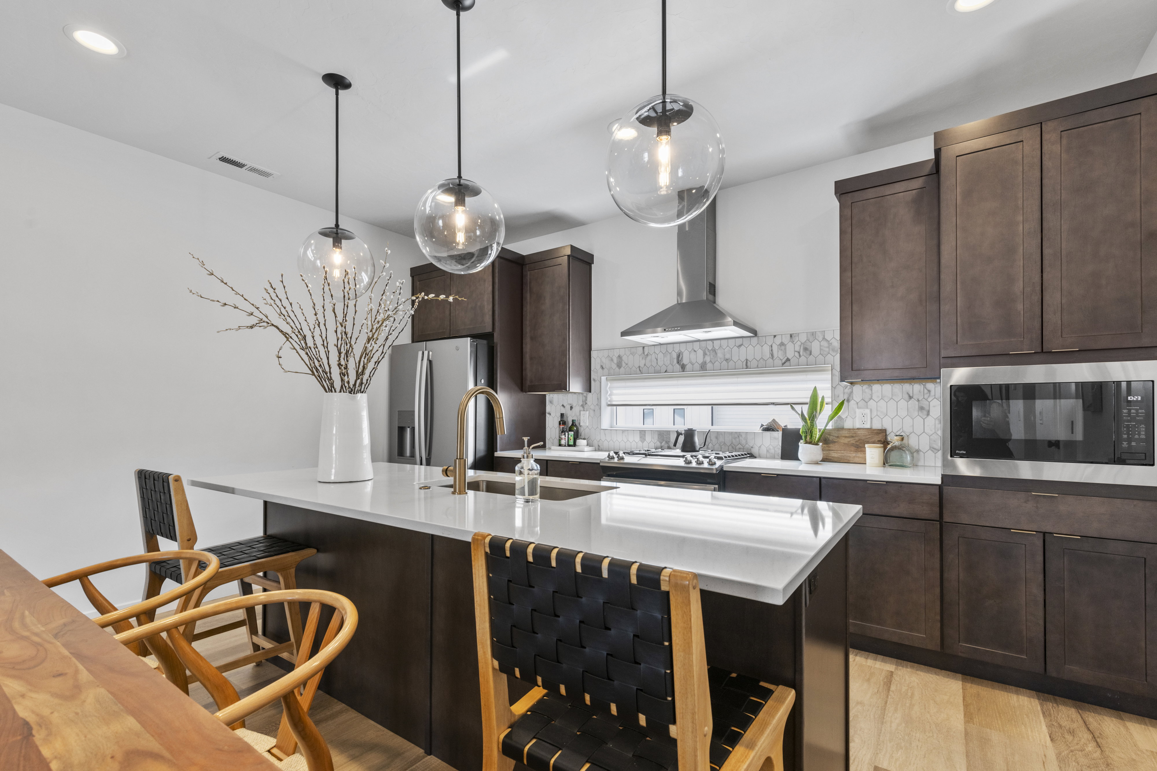 Contemporary kitchen featuring an expansive island with a light-toned countertop, dark wood-finish cabinetry, stainless steel appliances, and statement pendant lighting