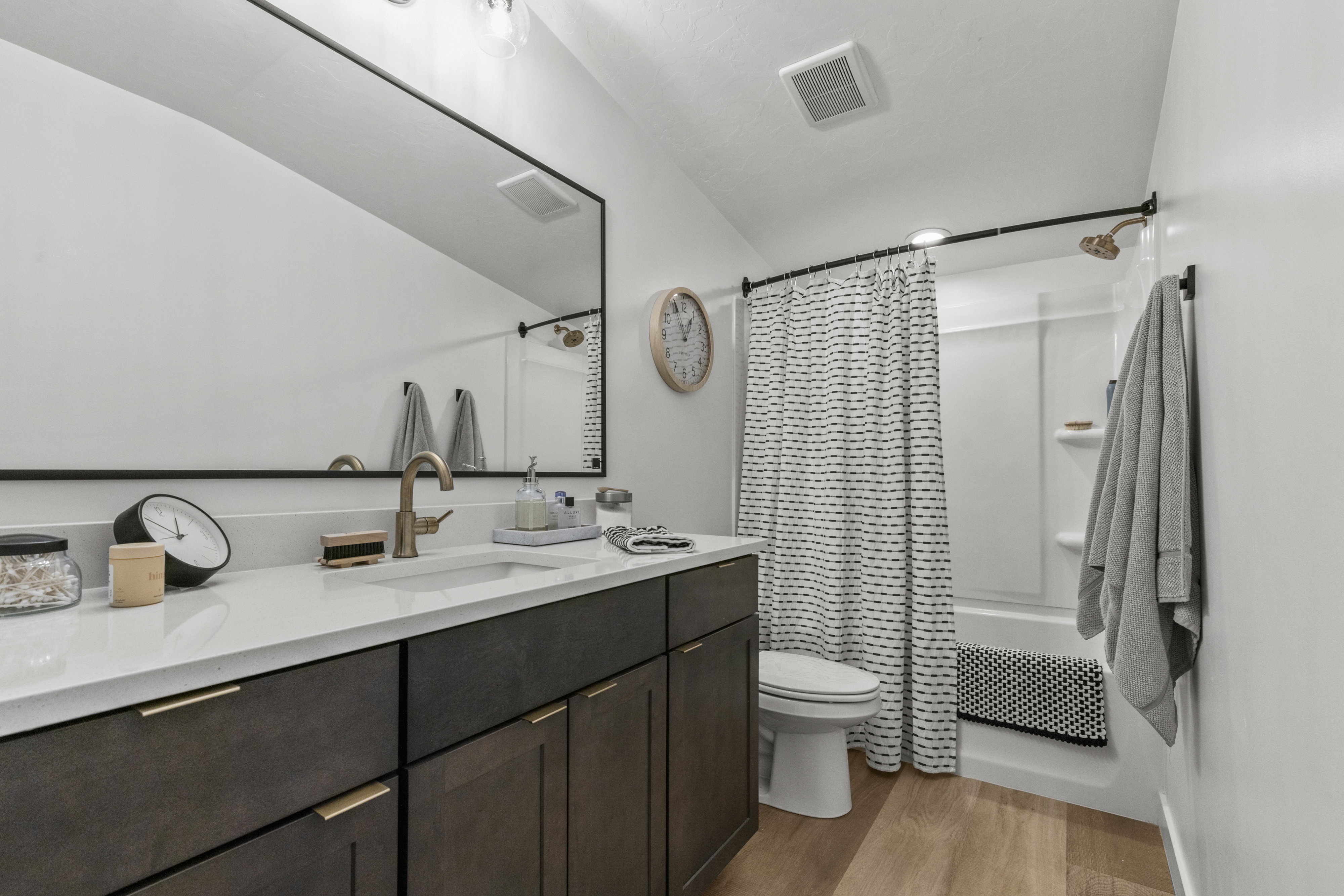 Contemporary bathroom featuring a dark wood-finish vanity with gold-tone hardware, a white countertop with an integrated sink, a rectangular framed mirror, and wood-finish flooring