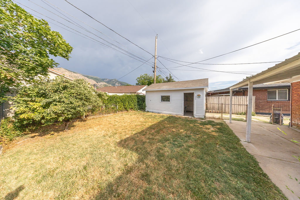 Expansive backyard featuring a detached storage shed, concrete patio with covered awning, and mature perimeter trees