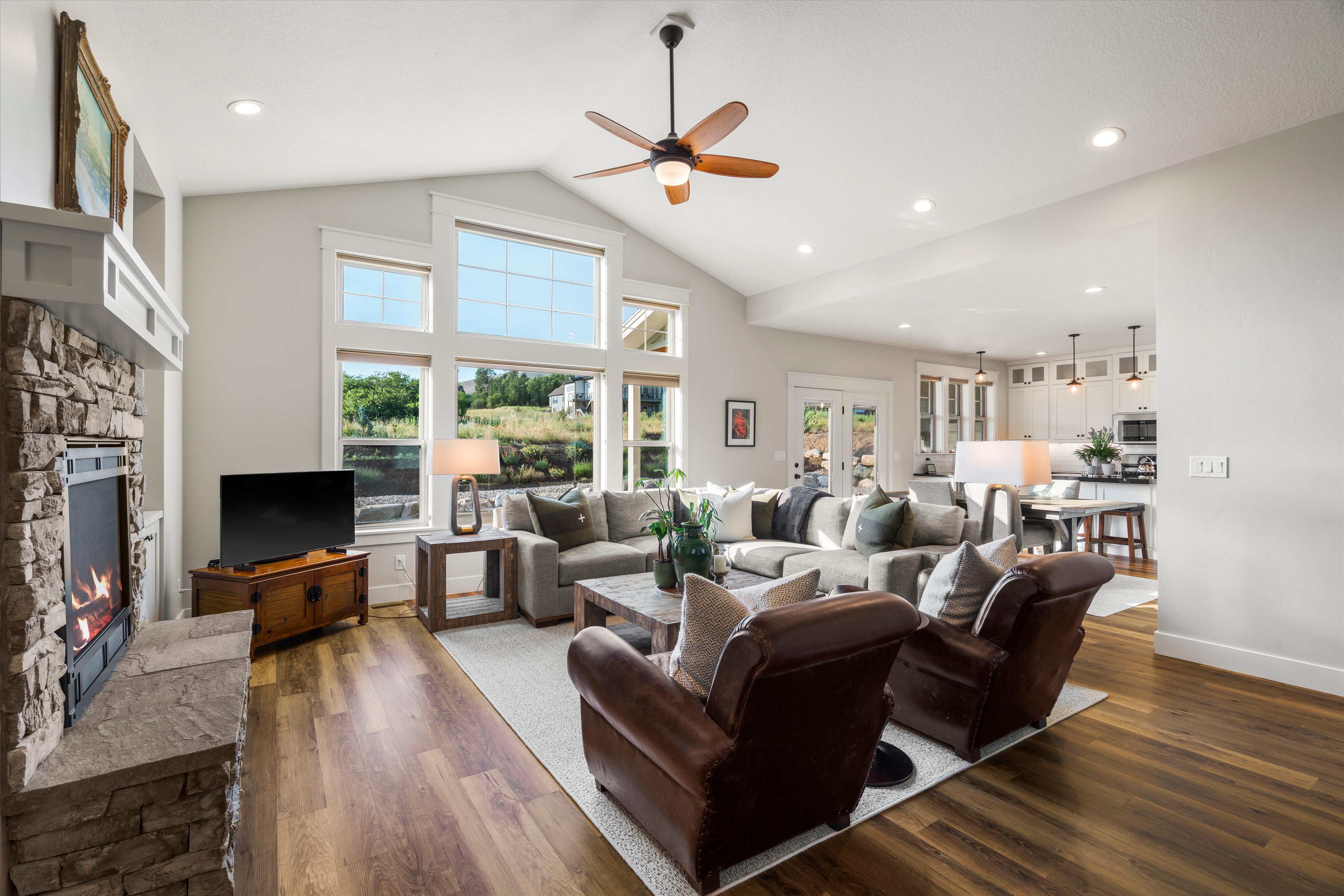 Vaulted living room with wood-finish flooring, a stone-surround fireplace, and expansive windows