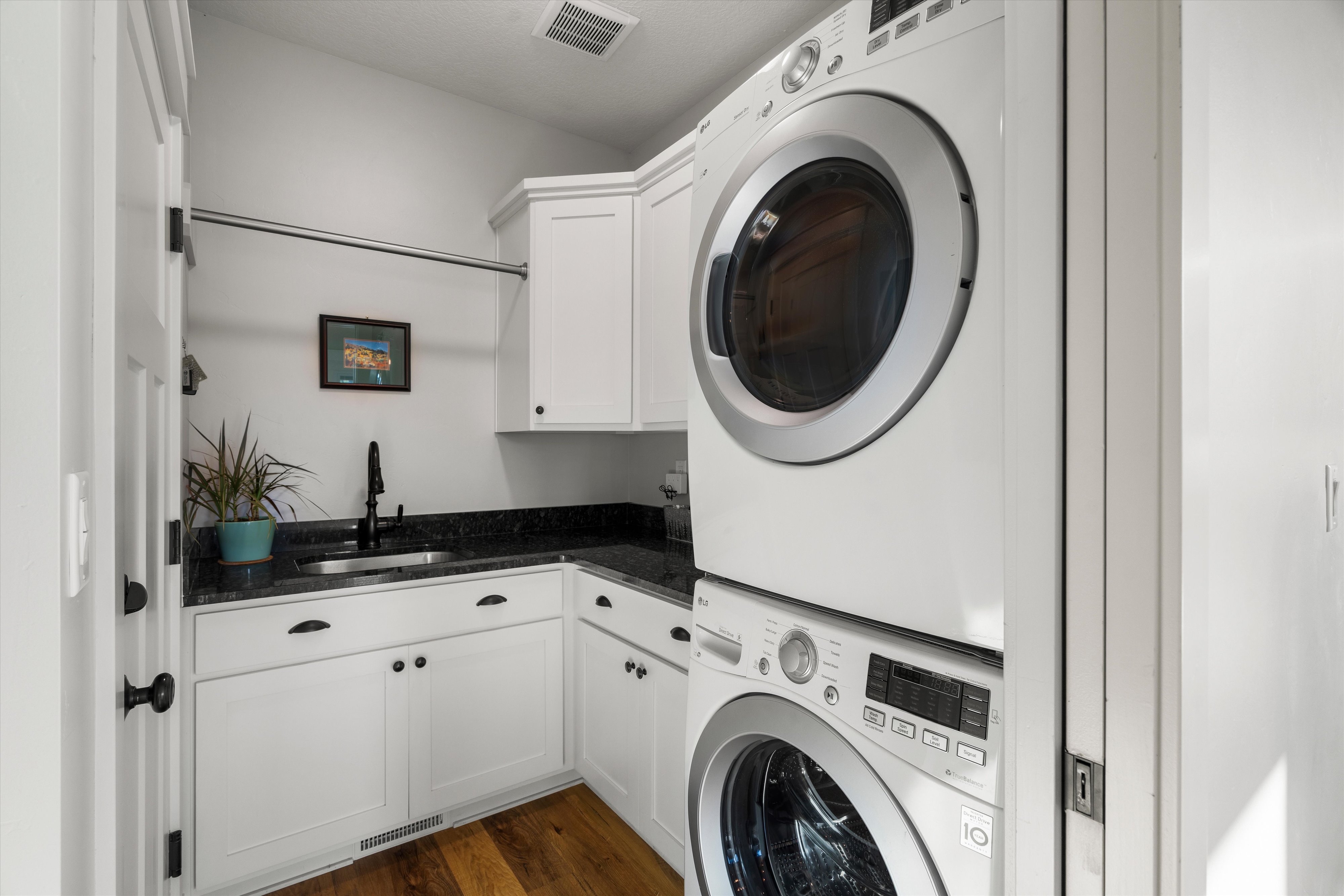 Dedicated laundry room featuring stacked appliances, ample white cabinetry, dark stone countertops, a stainless steel sink, and wood-finish flooring