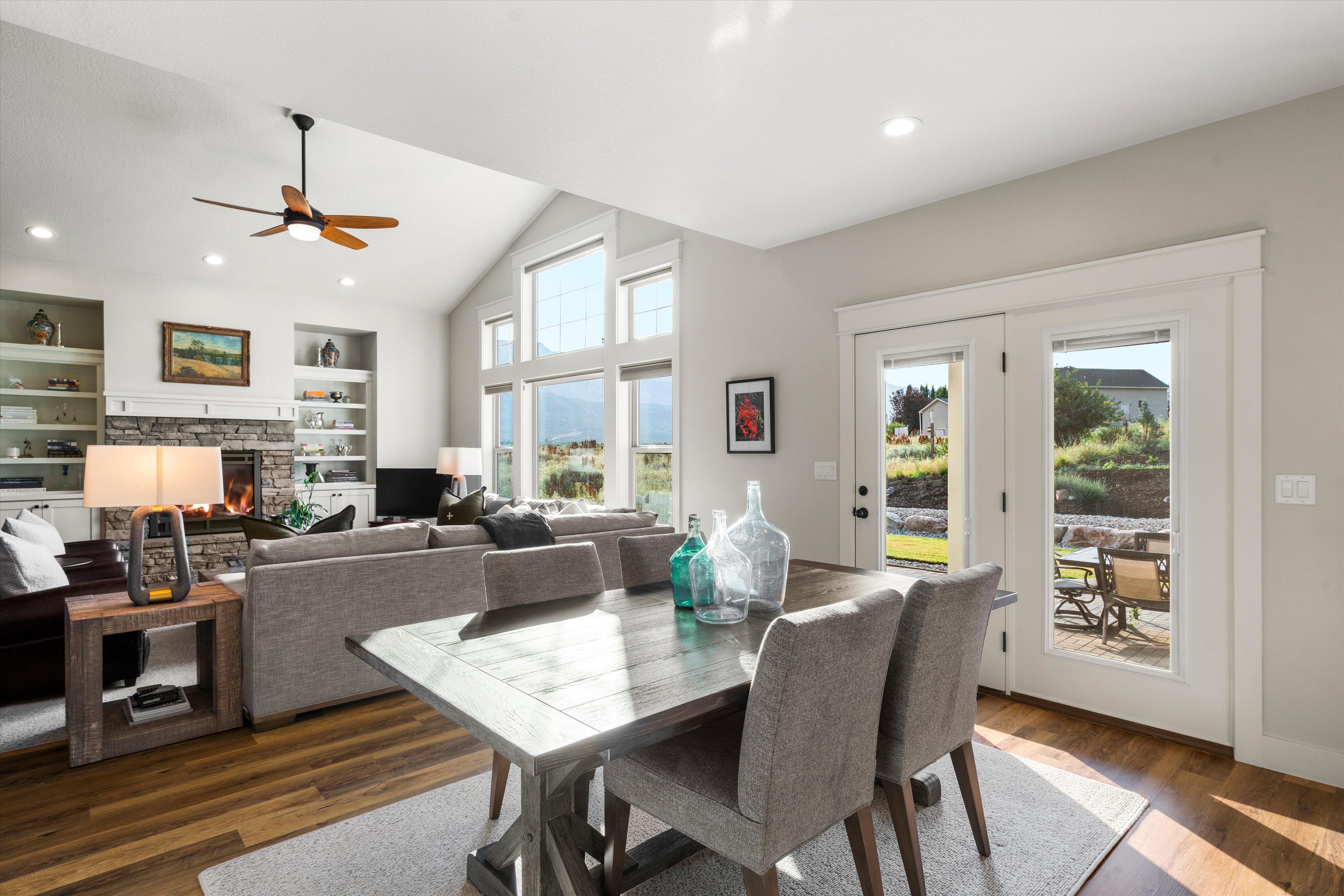Vaulted ceilings with a ceiling fan, a stone-front fireplace with built-in shelving, wood-finish flooring, and large windows with transom panels
