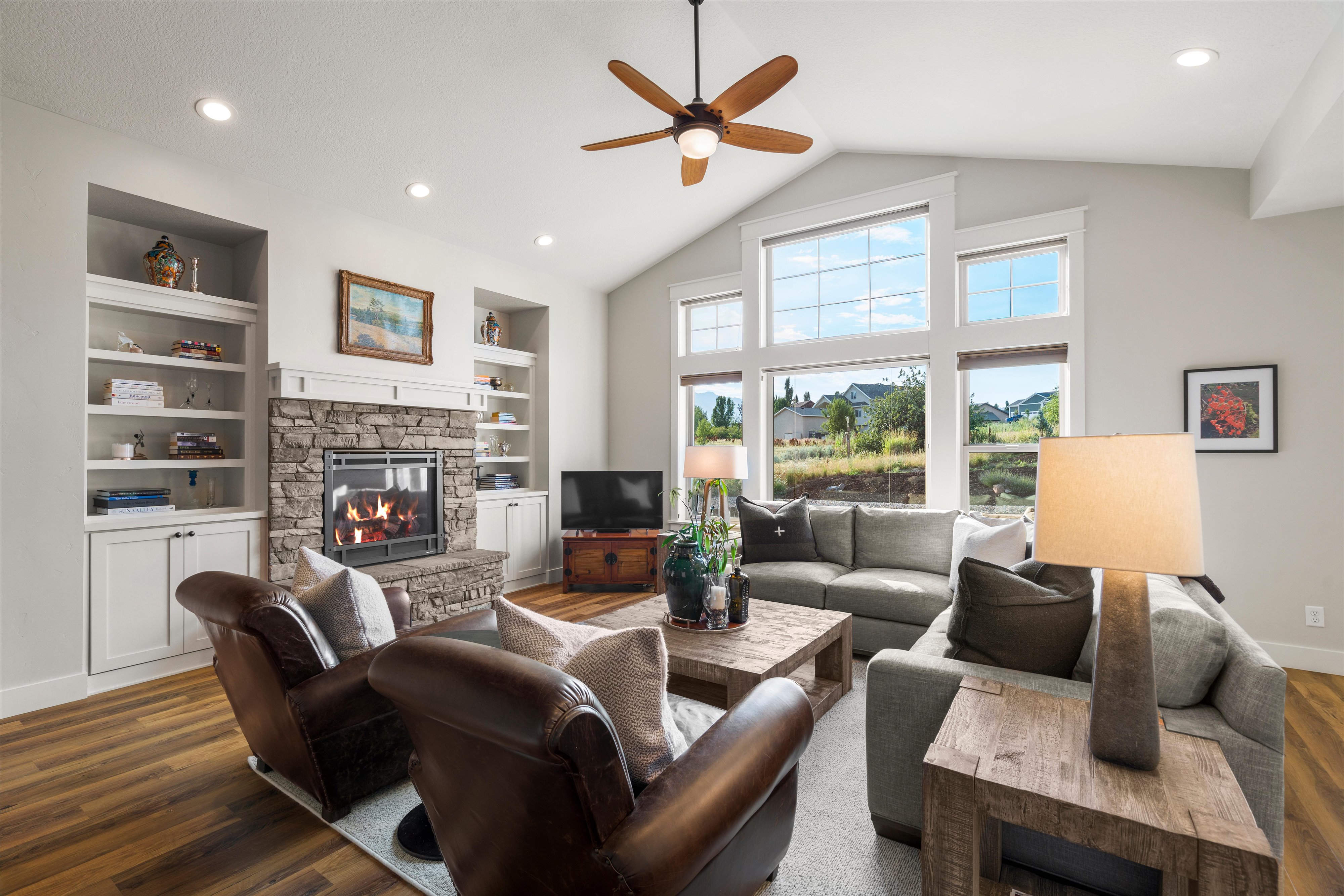 Vaulted ceiling living room featuring a stone-faced fireplace, integrated built-in shelving, wood-finish flooring, large picture windows, and recessed lighting