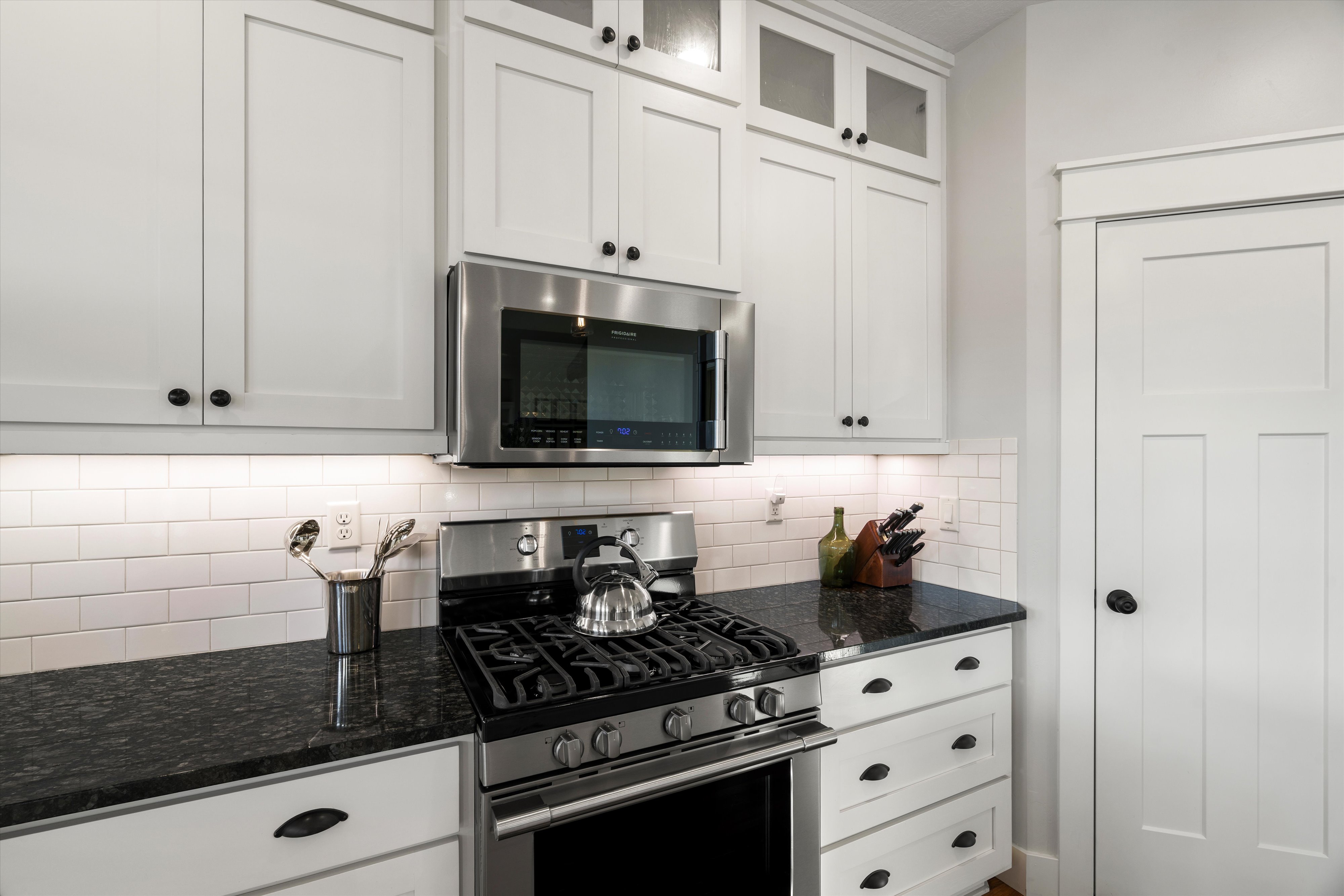 White shaker-style cabinetry with black hardware, stainless steel appliances, dark stone countertops, and white subway tile backsplash