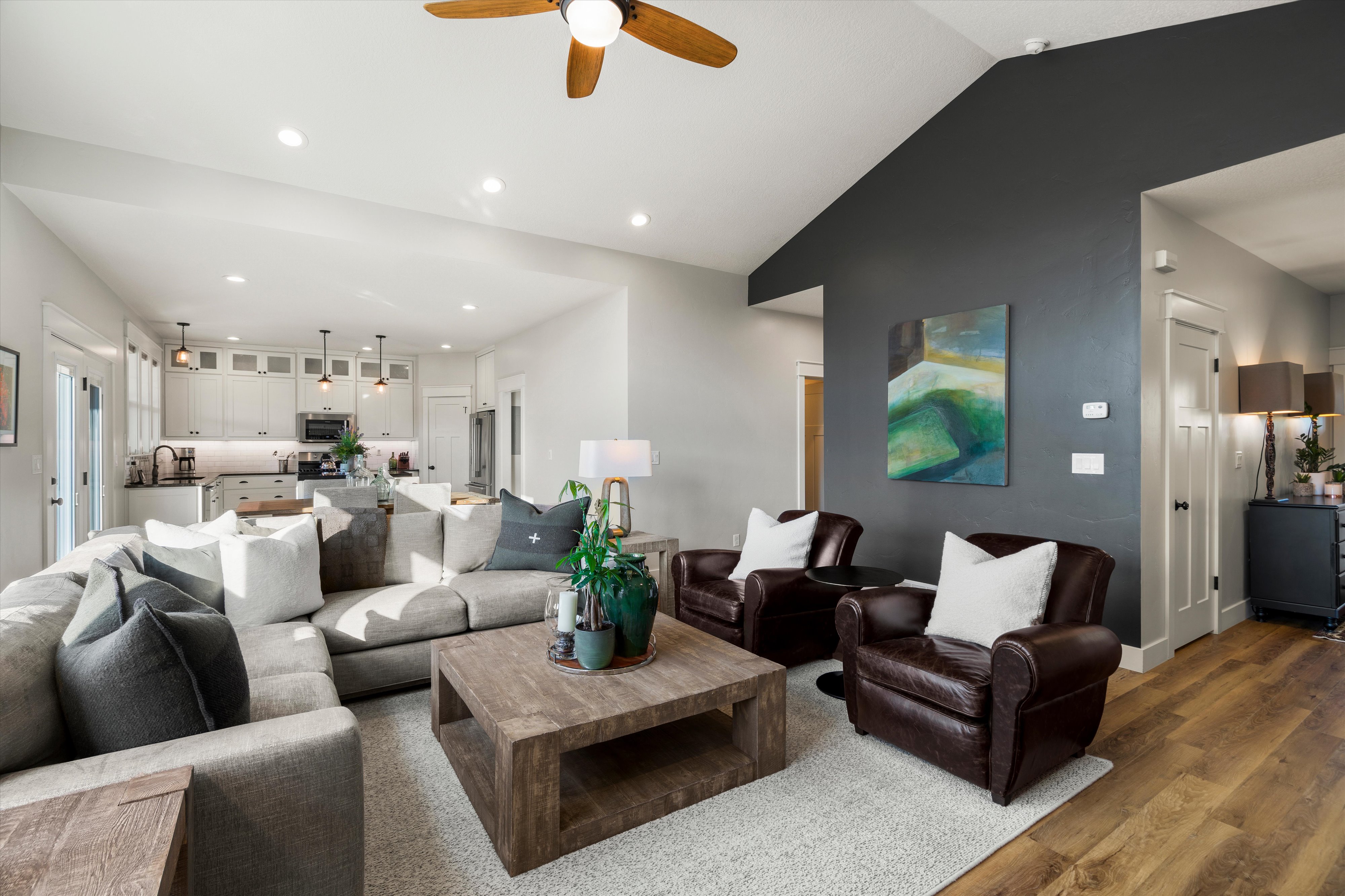 Spacious living area featuring a vaulted ceiling with recessed lighting and a ceiling fan, wood-finish flooring, a contemporary accent wall, and a bright kitchen visible in the background