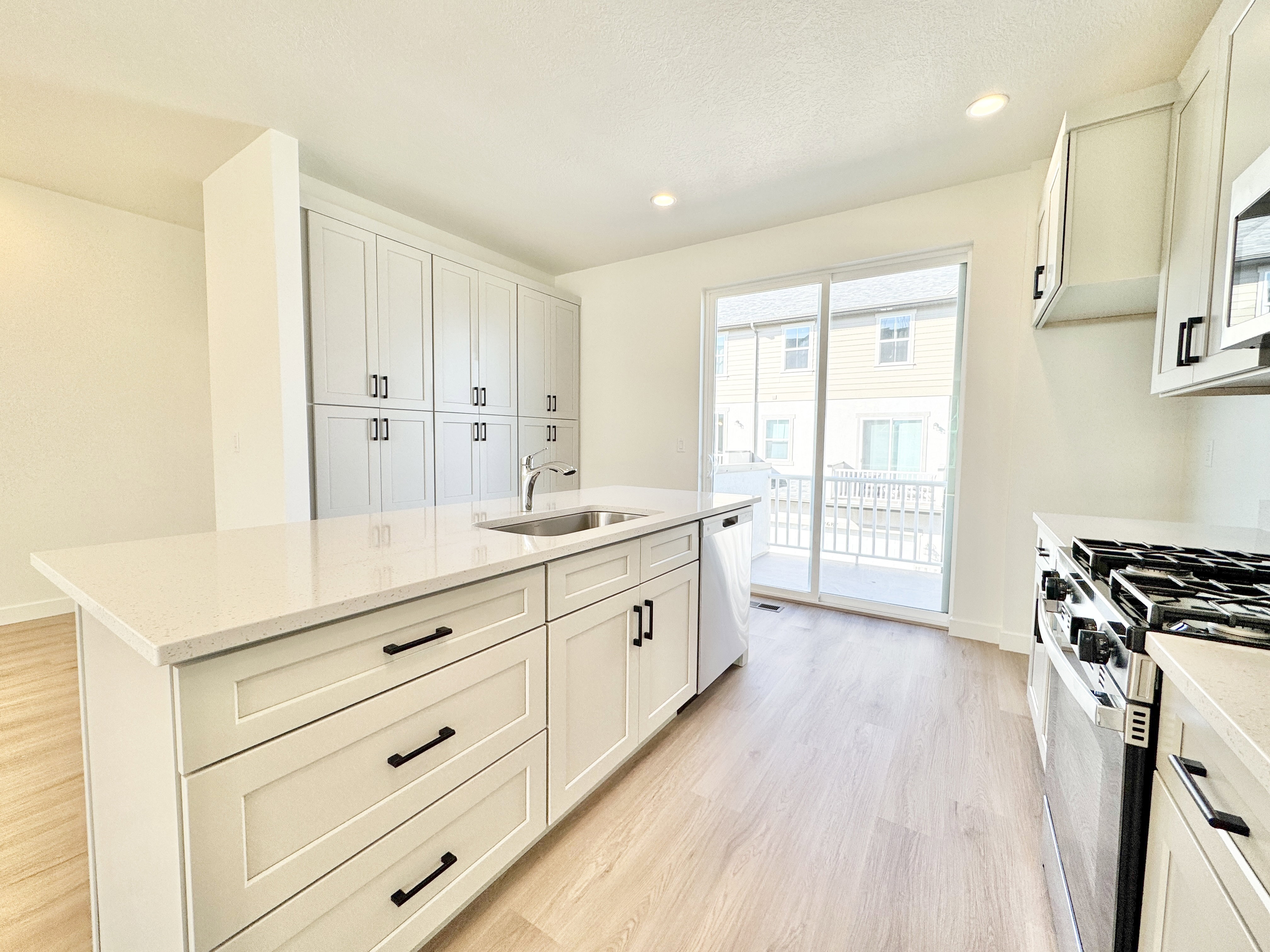 Modern kitchen featuring an island with a stainless steel sink, light wood-finish flooring, white cabinetry with dark hardware, recessed lighting, and a sliding glass door leading to a balcony