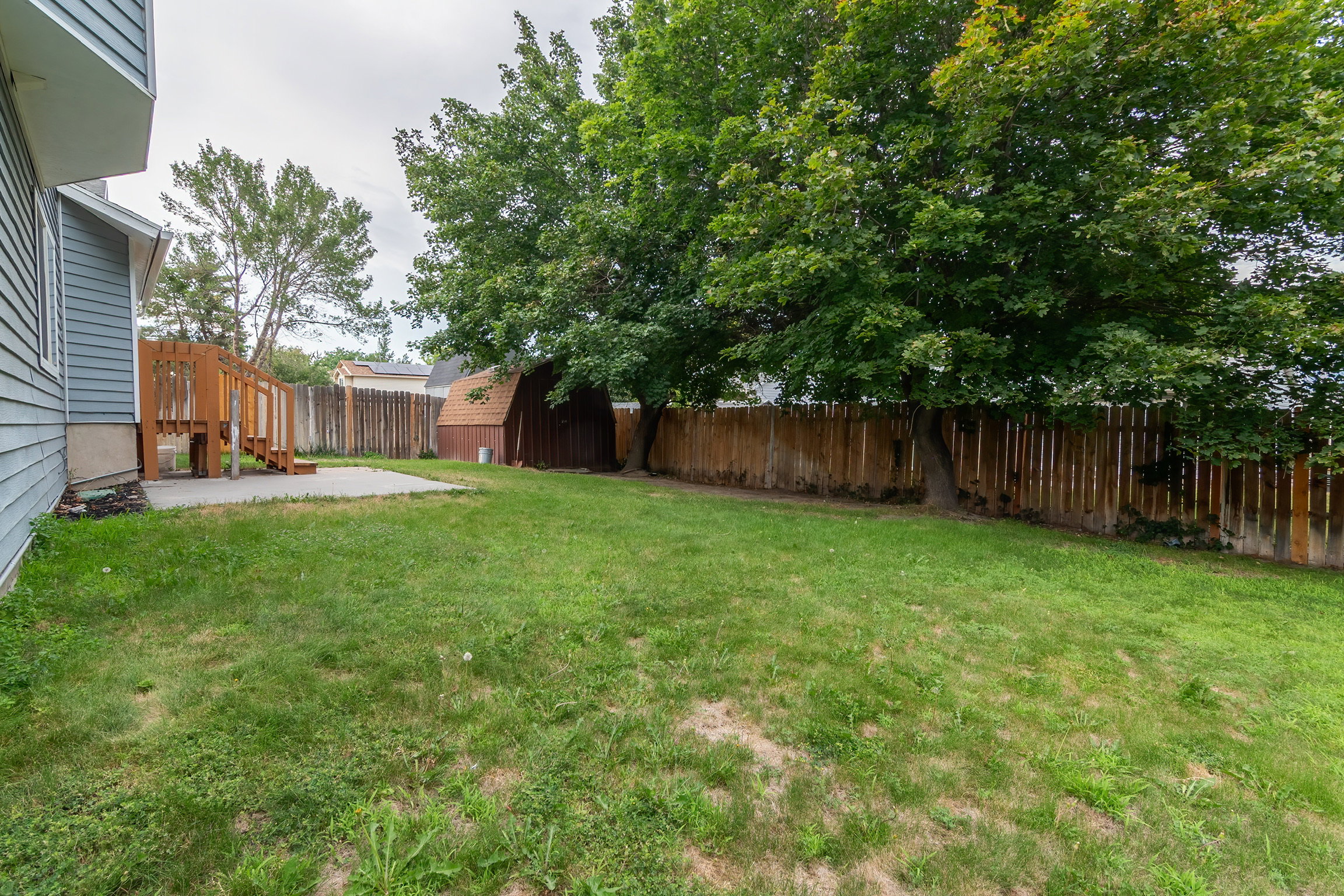 Spacious backyard featuring a large concrete patio, wood deck with railing, mature trees, and a privacy fence