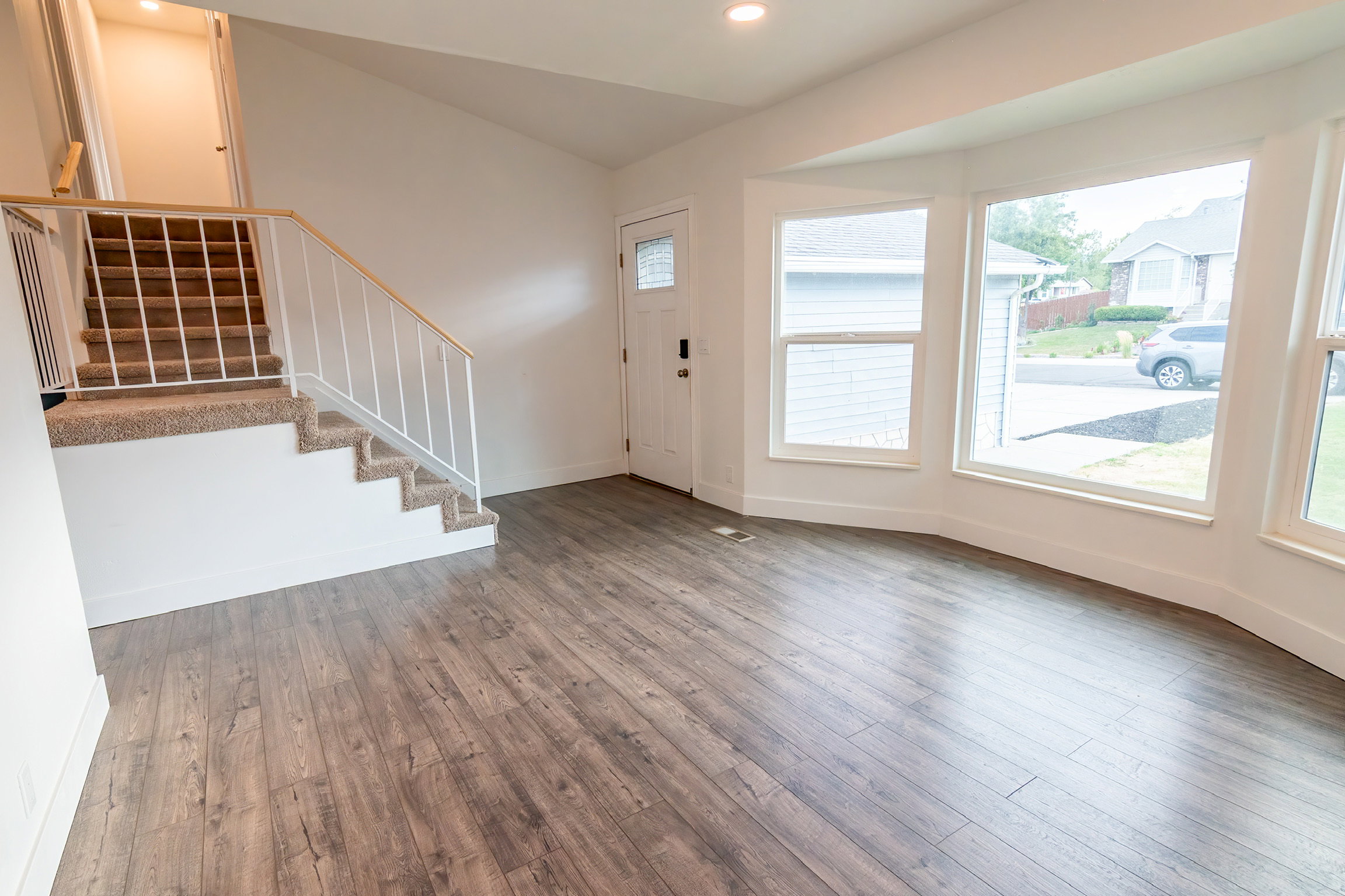 Spacious living area featuring wood-finish flooring, a bay window with multiple panes, and a carpeted staircase with a white metal railing