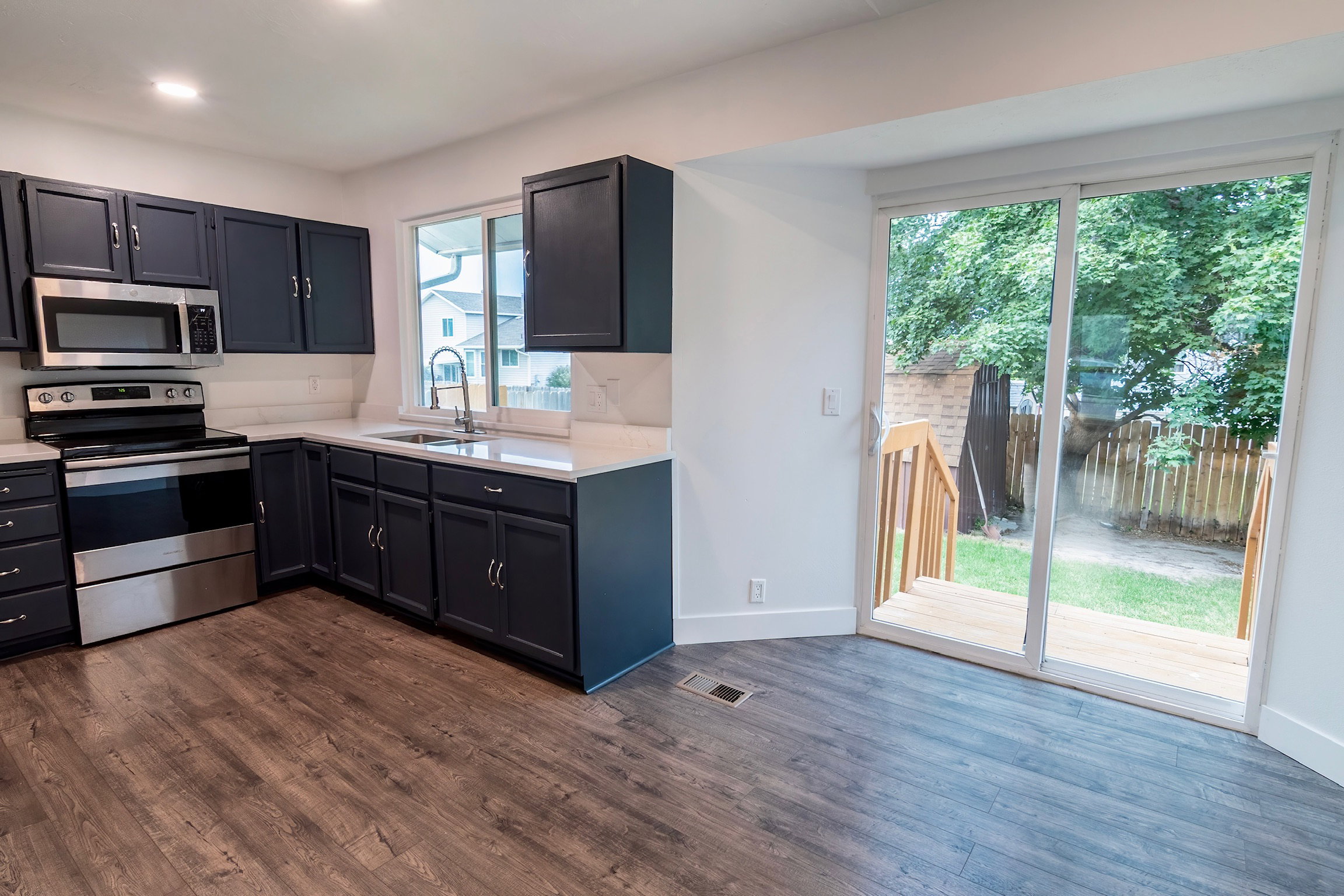 Kitchen featuring dark cabinetry, stainless steel appliances, a white countertop, wood-finish flooring, and a sliding glass door leading to a wooden deck and enclosed yard