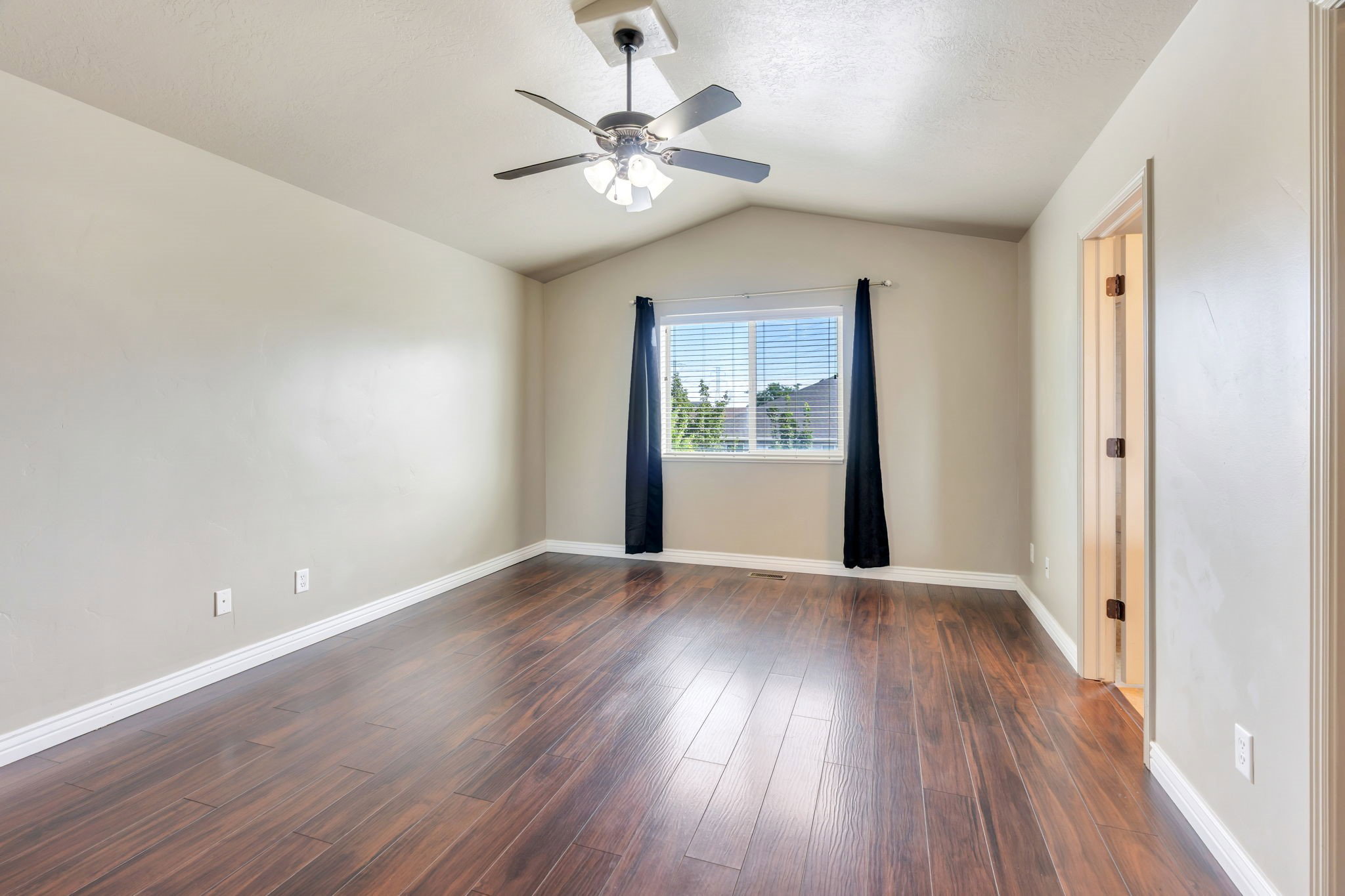 Spacious room featuring vaulted ceilings, wood-finish flooring, a ceiling fan with integrated lighting, and a large window with blinds