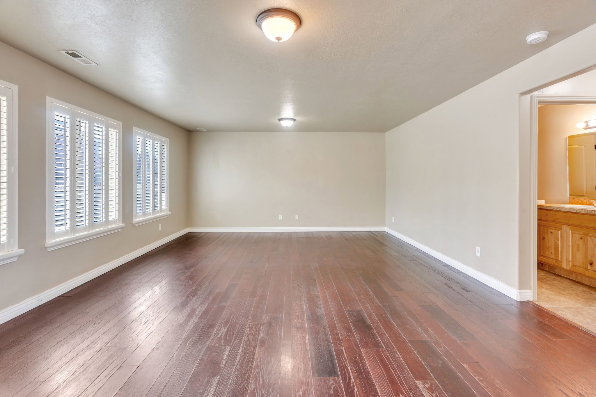 Spacious room featuring rich wood-finish flooring, multiple windows with white plantation shutters, and neutral wall tones