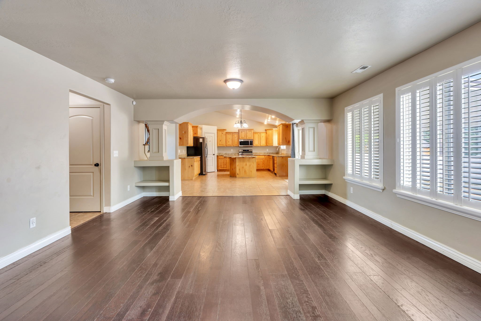 Spacious room featuring wood-finish flooring, white plantation shutters, and an arched entryway