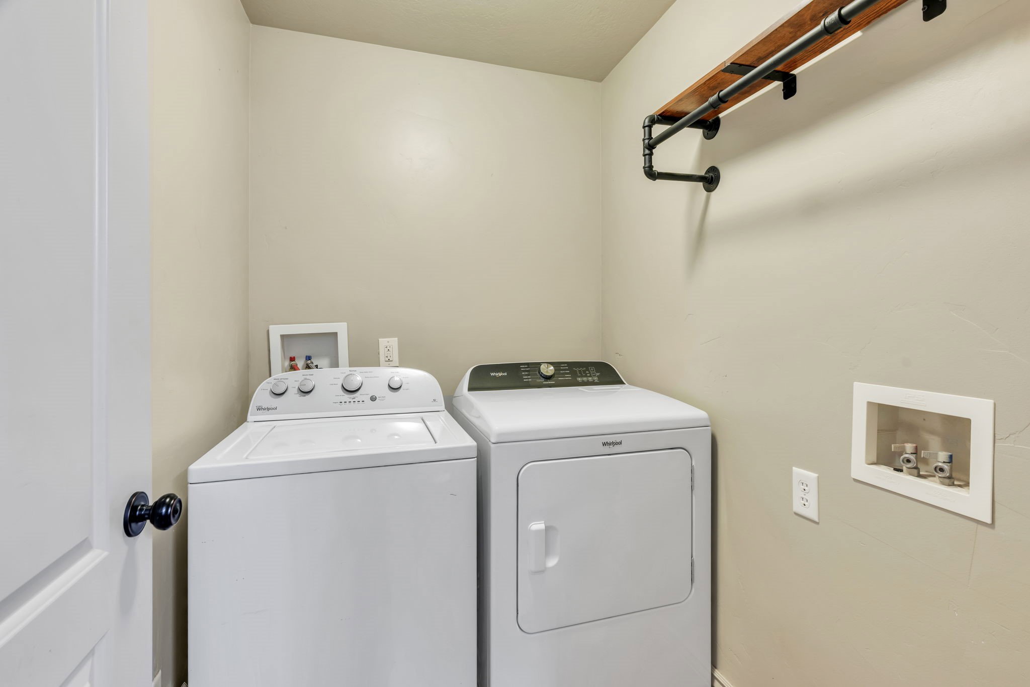 Dedicated laundry space featuring a wood-finish shelf with industrial-style piping, neutral wall paint, and a white interior door with a black doorknob
