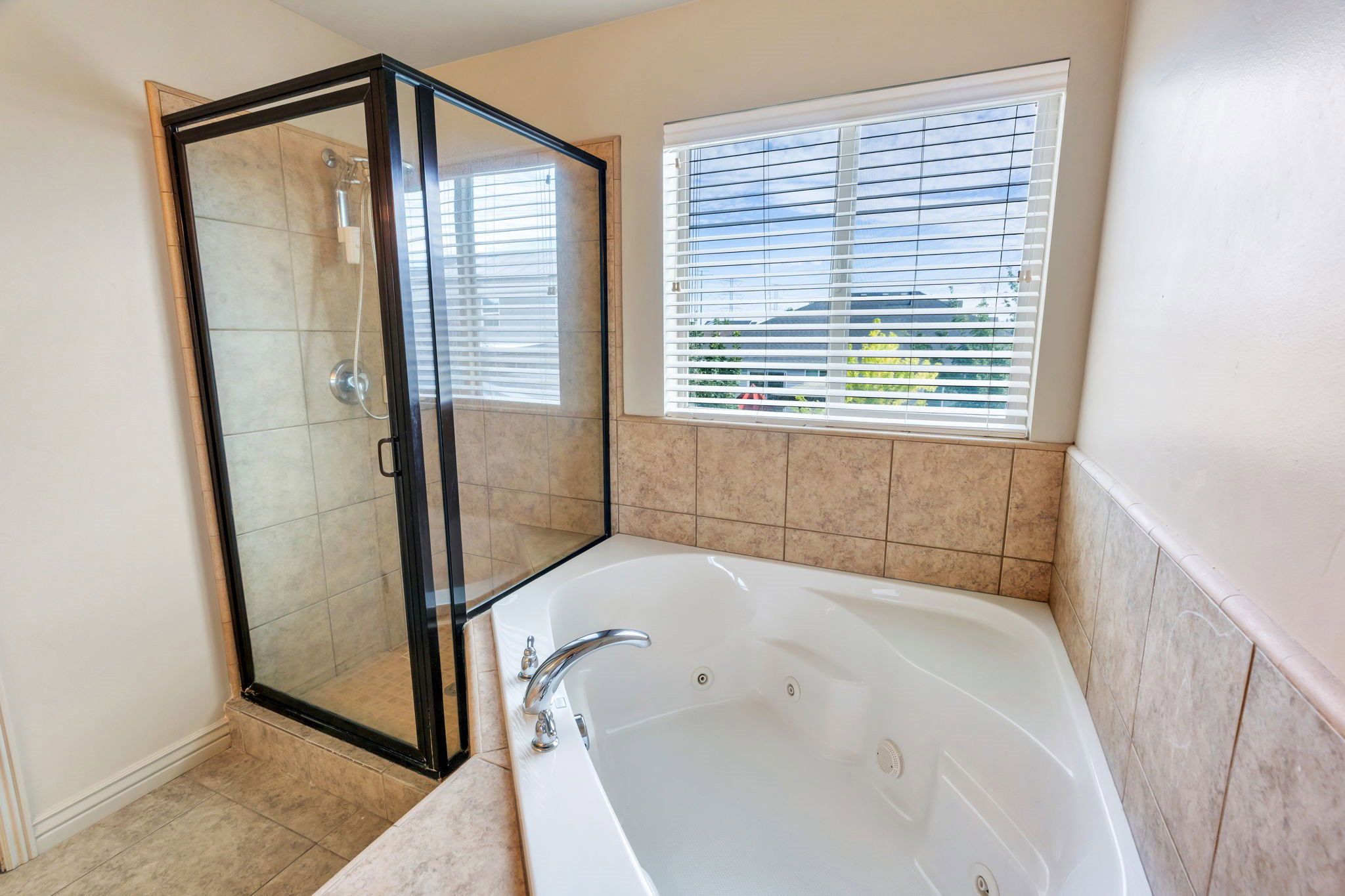 Bathroom featuring a corner jetted tub with chrome fixtures, a separate glass-enclosed shower with tile surround, and a window with blinds